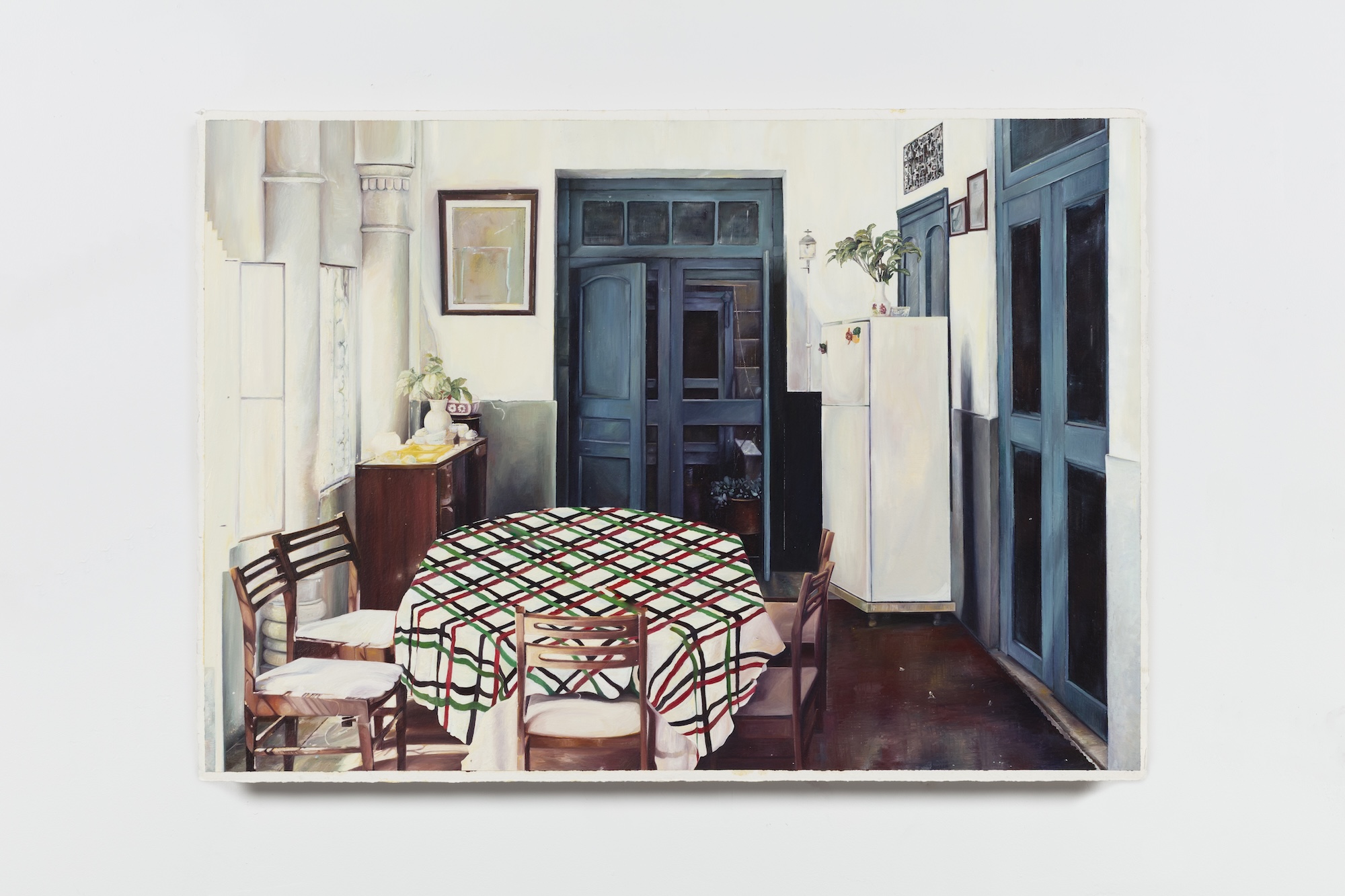 Realist painting of a Mediterranean-style dining room interior, showing a round table with a red, green, and black plaid tablecloth surrounded by wooden chairs, a white refrigerator, and tall blue-paneled doors.