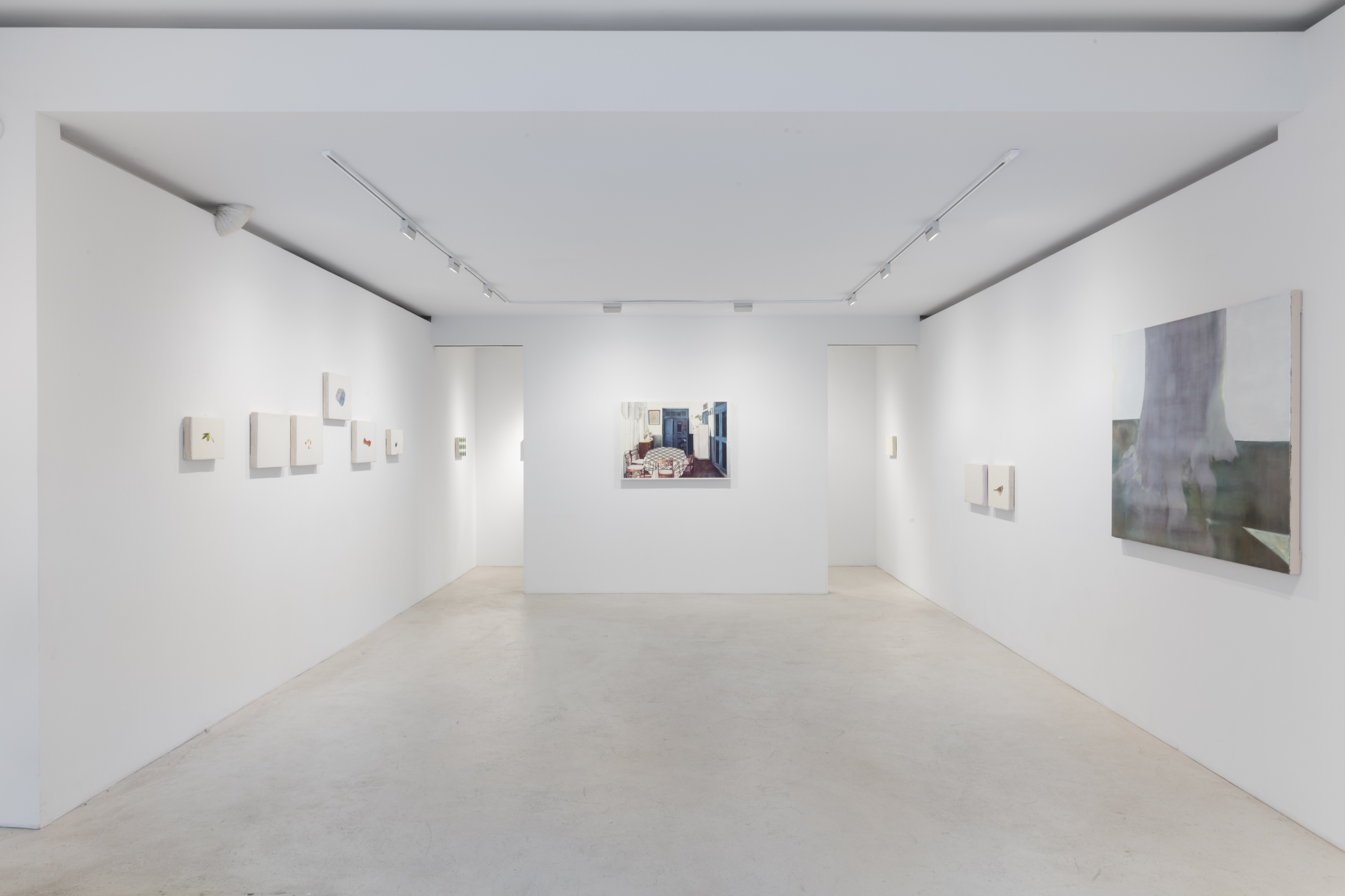 Installation view of a gallery exhibition with white walls, showing a cluster of small intimate paintings on the left, a larger interior scene centered on a freestanding wall, and an abstract figurative painting on the right.