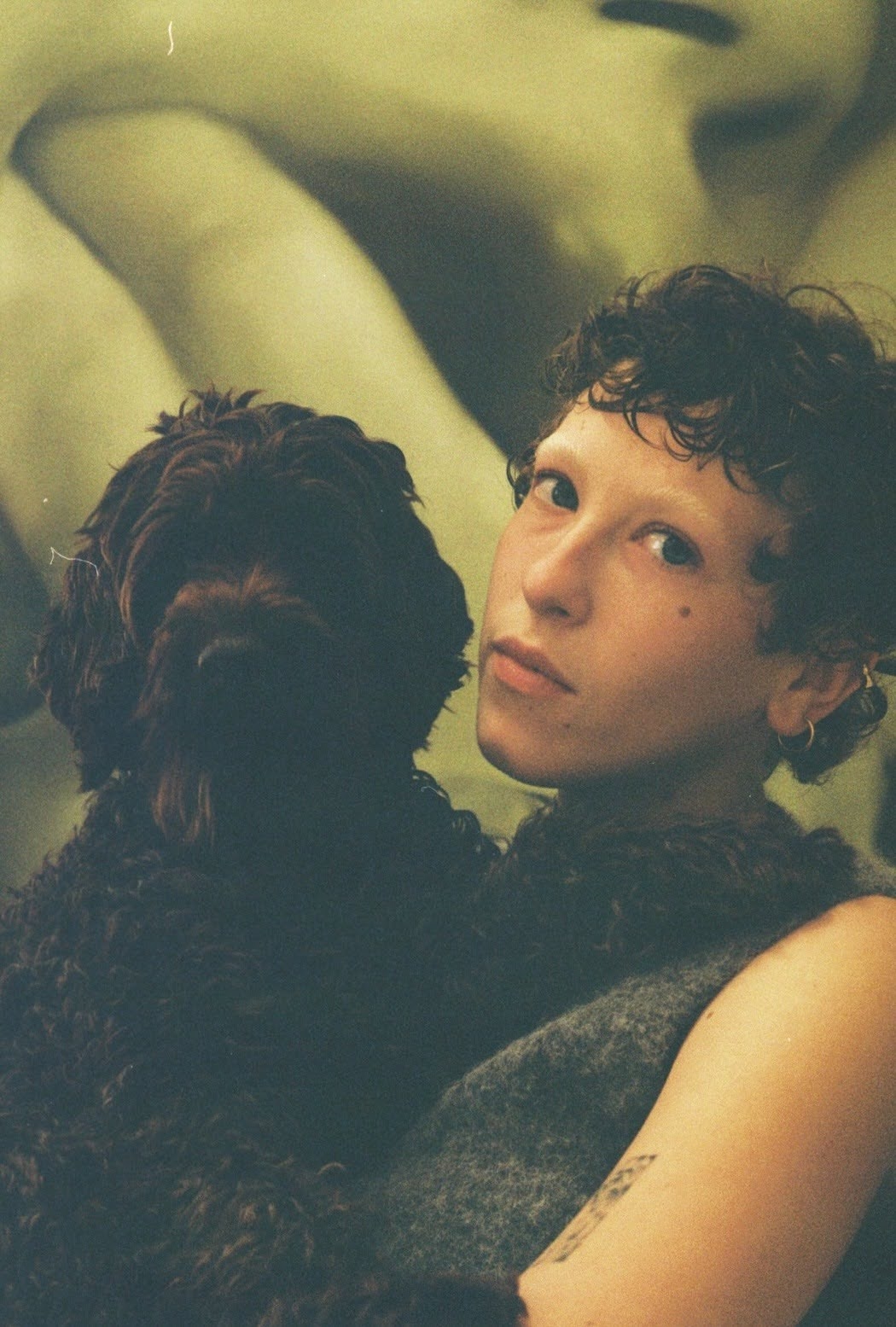 Color photograph of a young woman with curly brown hair looking over her shoulder at the camera, a dark curly-haired dog beside her, against a green-tinted blurred background.