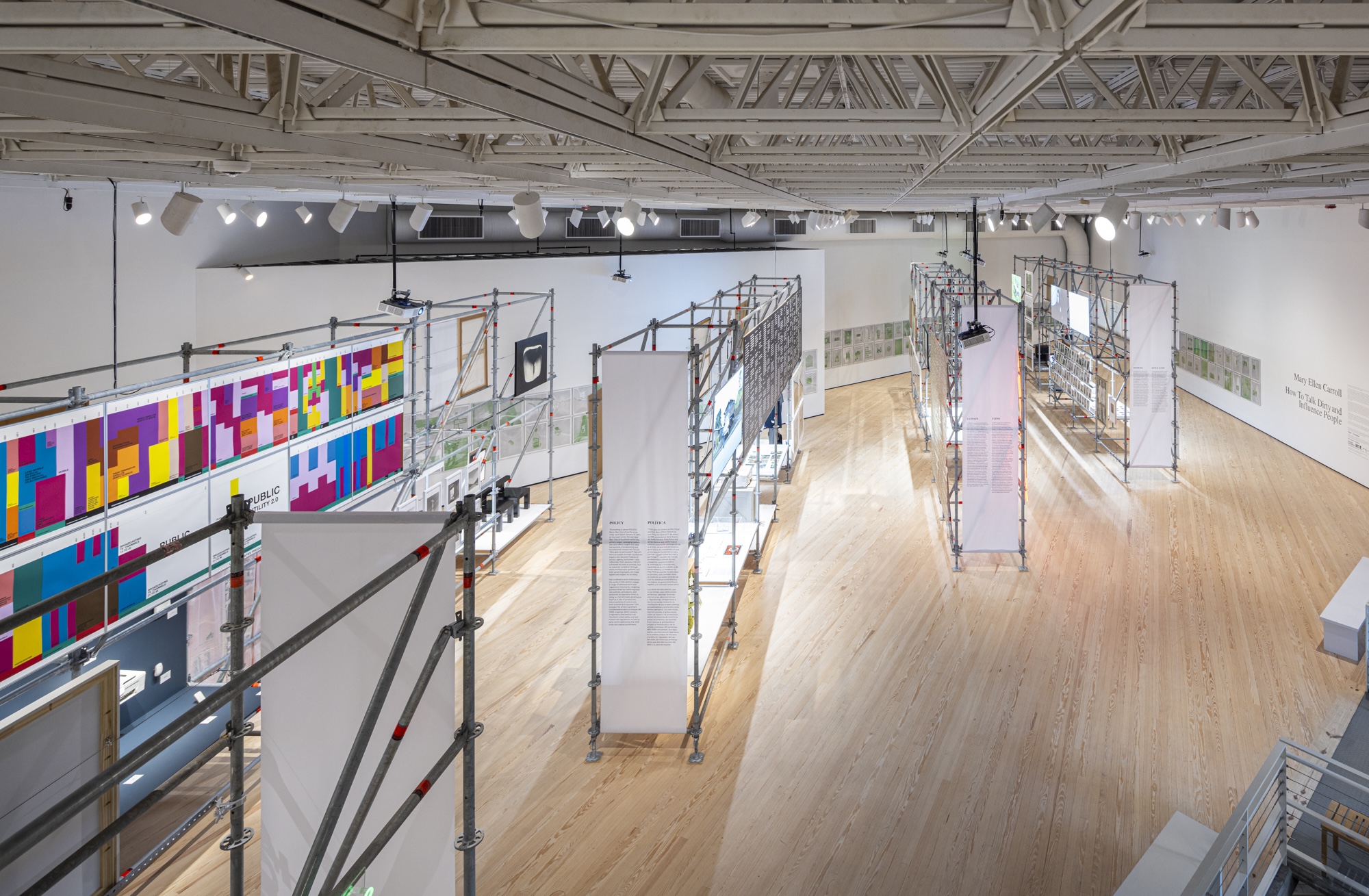 Elevated view of a museum gallery with scaffolding display structures holding colorful charts, framed artworks, and hanging text panels, part of an exhibition titled "How To Talk Dirty and Influence People."
