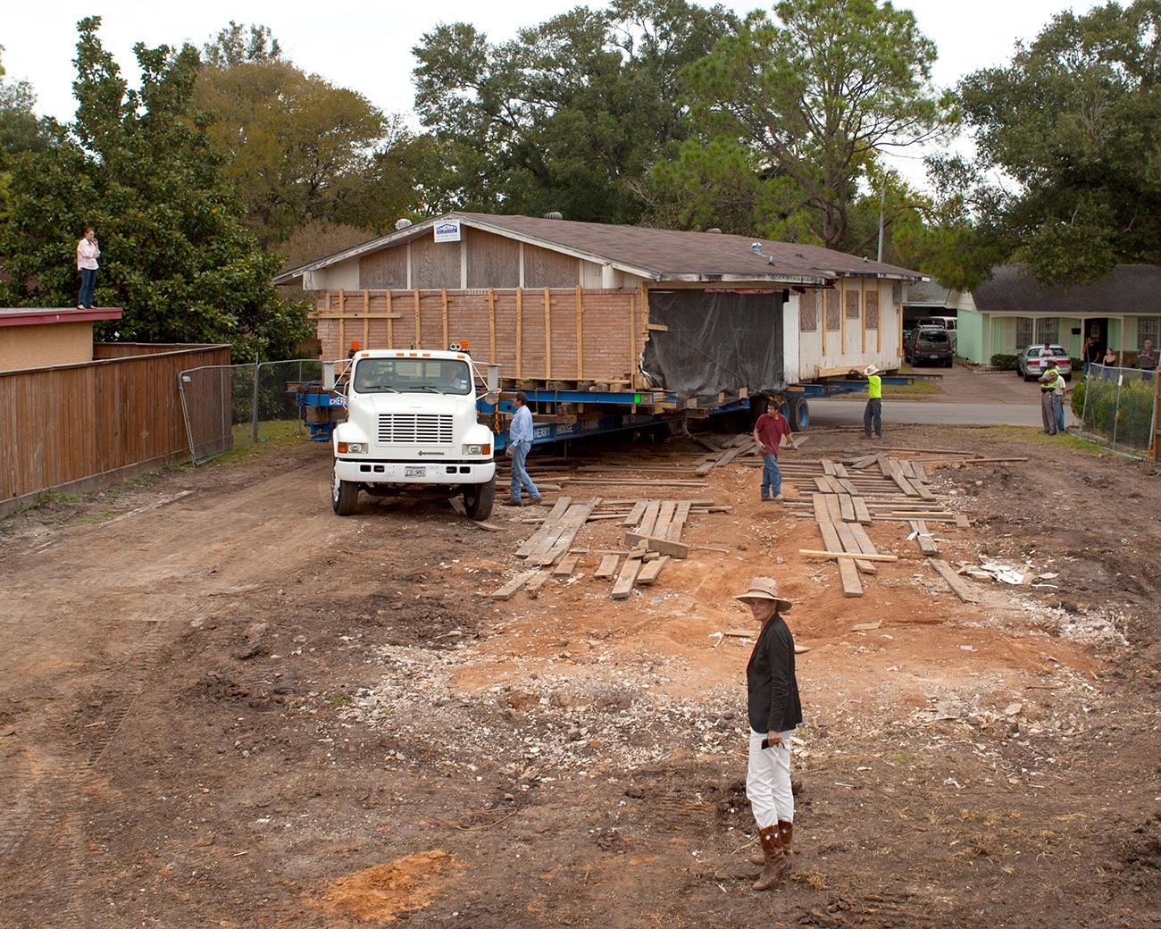 A single-story house being relocated on a flatbed truck trailer, with workers and onlookers standing in a dirt yard, including a woman in a hat and white pants in the foreground.