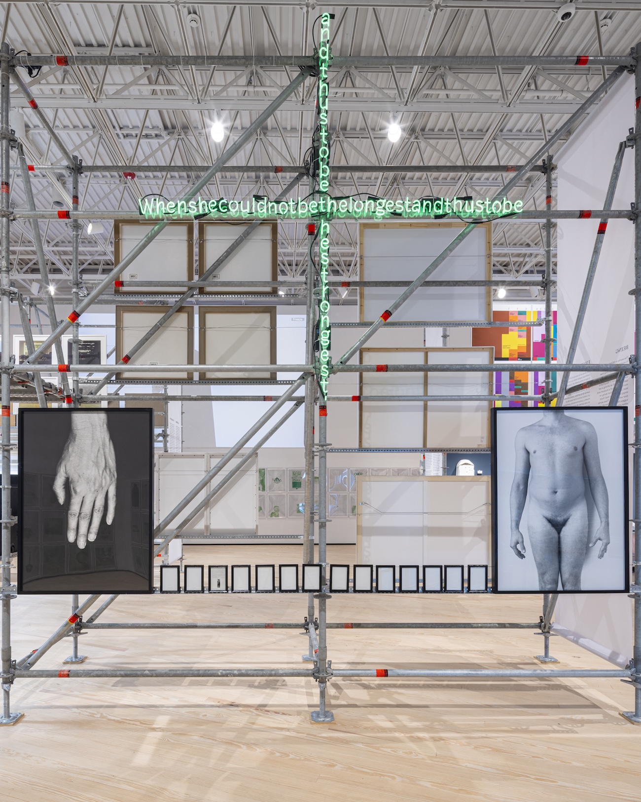 Gallery installation with scaffolding structure holding a black-and-white photo of a hand, a nude figure photo, and a row of small framed images, beneath a green neon text sculpture reading a cursive-style sentence.