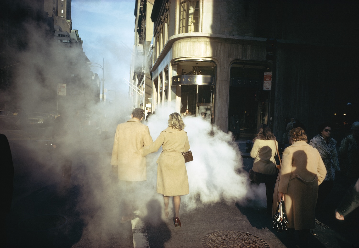 Two women in matching camel coats walk arm-in-arm through steam rising from a Manhattan sidewalk grate, near a Gucci storefront on a sunny day.