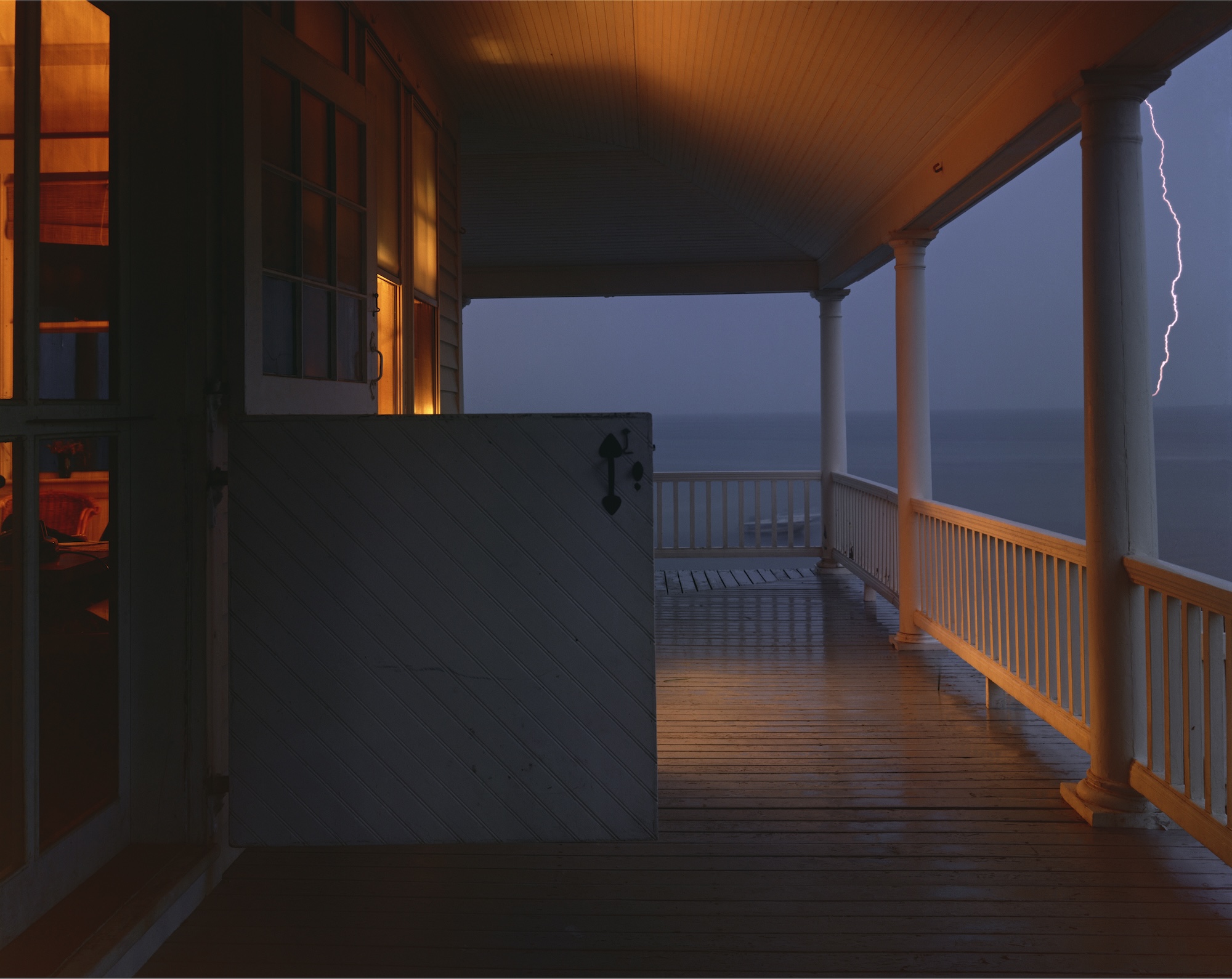 A wraparound porch at dusk overlooks the ocean, warm interior light spilling out as a bolt of lightning streaks across the darkening sky.