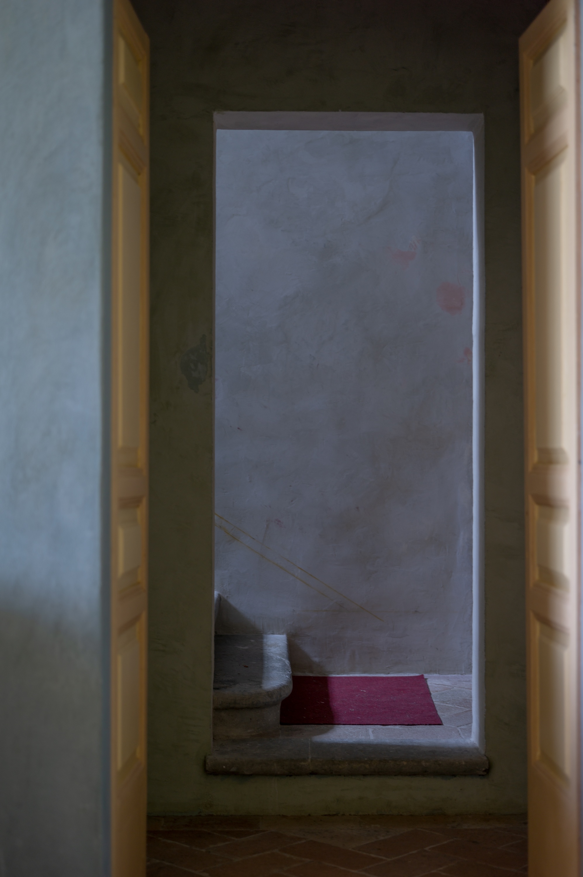 A dim hallway frames a bright, sparsely furnished room beyond, with a magenta mat on the floor and a pale armchair against a stained plaster wall.