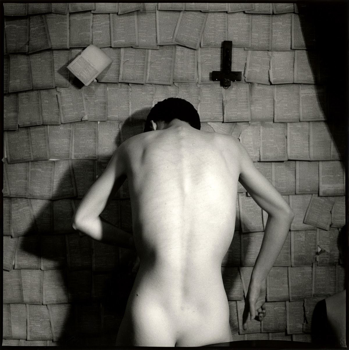 Black-and-white photo of a nude figure seen from behind, standing before a wall covered in torn book pages, with a small wooden cross and an open book mounted above.