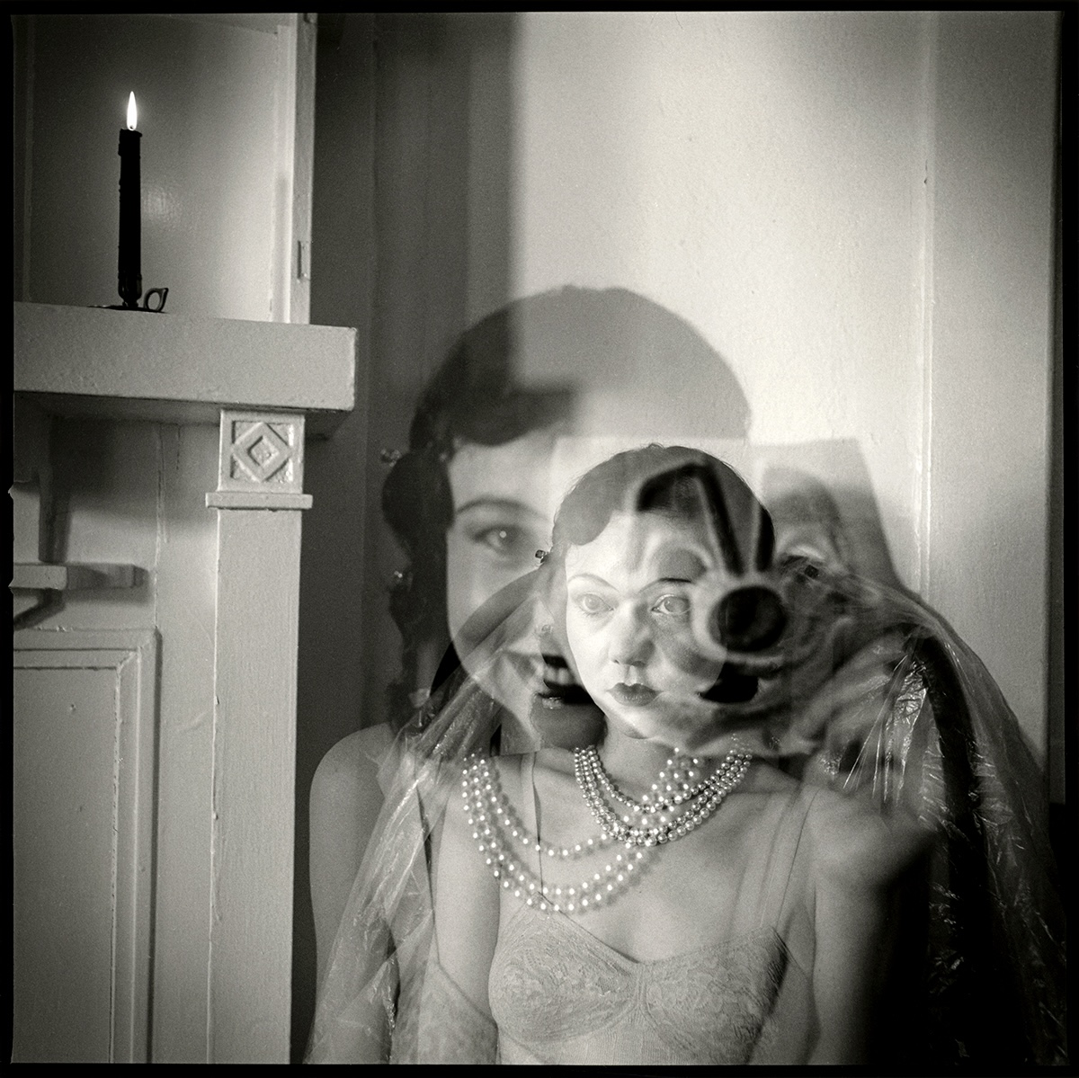 Black-and-white double-exposure portrait of a woman in a lace slip and layered pearl necklaces, her face blurred by movement, with a lit candle on a mantel beside her.
