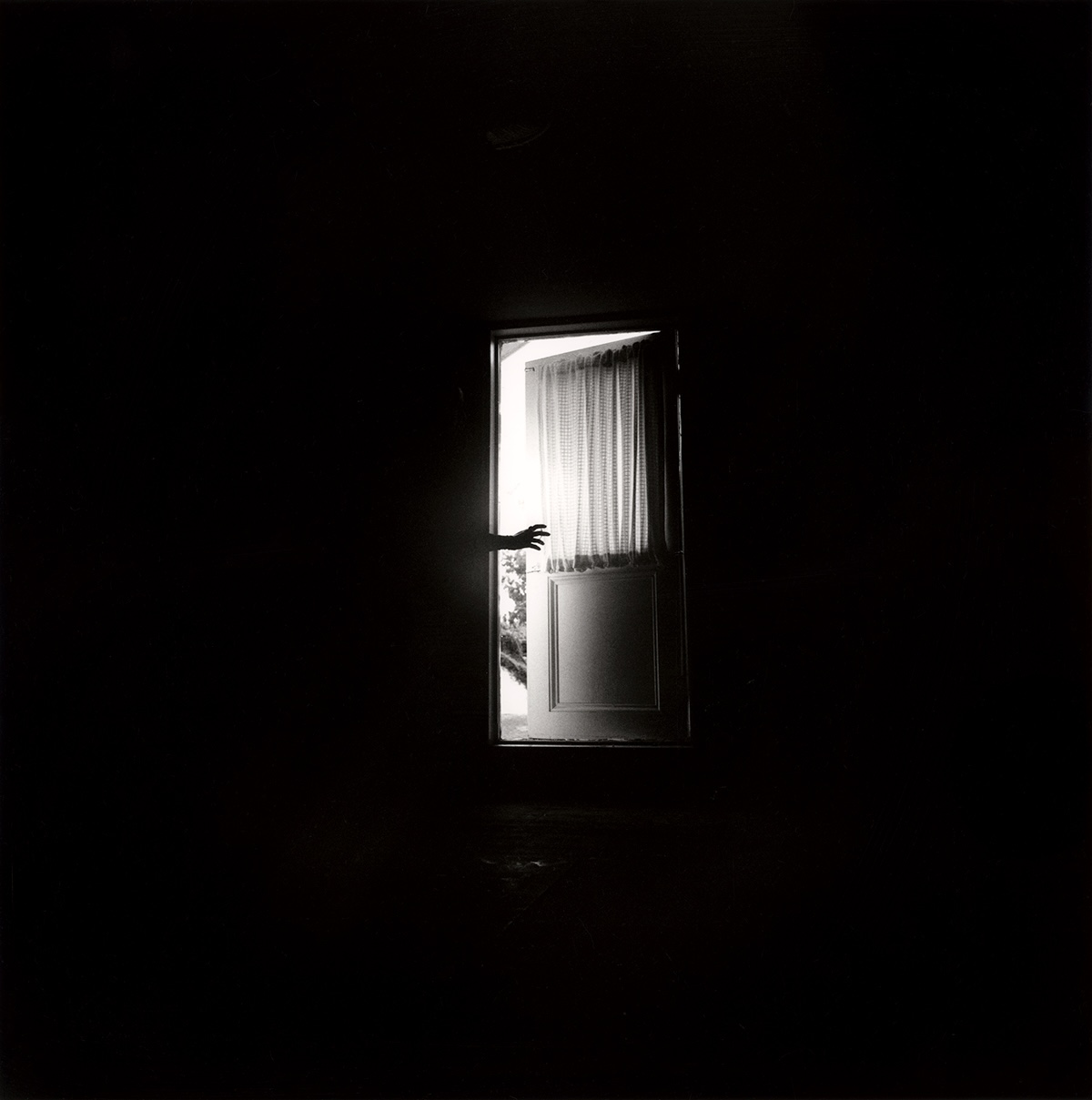 Black-and-white photo of a dark, nearly empty room with a single lit doorway in the background; a hand reaches into the doorway beside a curtain, the rest of the room in deep shadow.