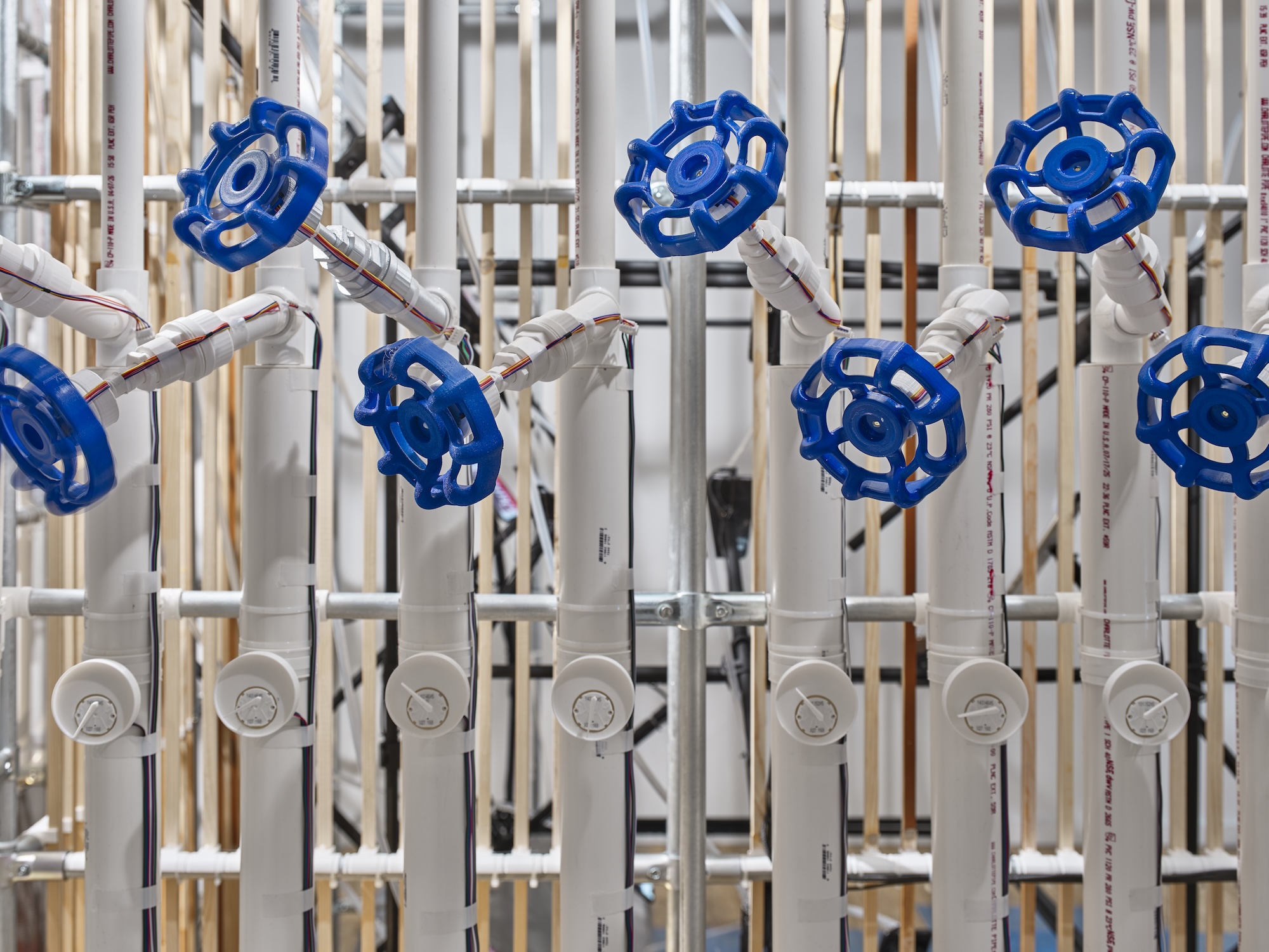 Close-up of blue valve handles mounted on white PVC pipes, each fitted with wiring and small gauges below, arranged in a row against a backdrop of wooden dowels and metal poles.