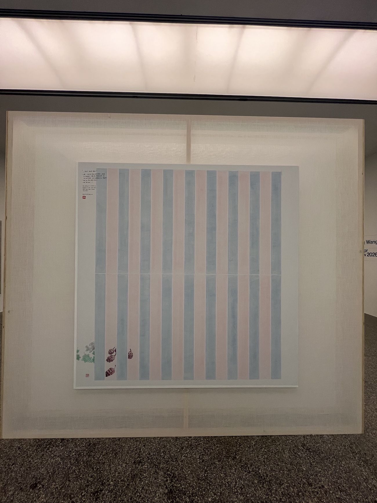 Gallery installation view of a striped painting by Evelyn Taocheng Wang hanging on a fabric-covered wall, featuring alternating pale blue and pink vertical bands with handwritten text and small painted botanical motifs in the lower left corner.
