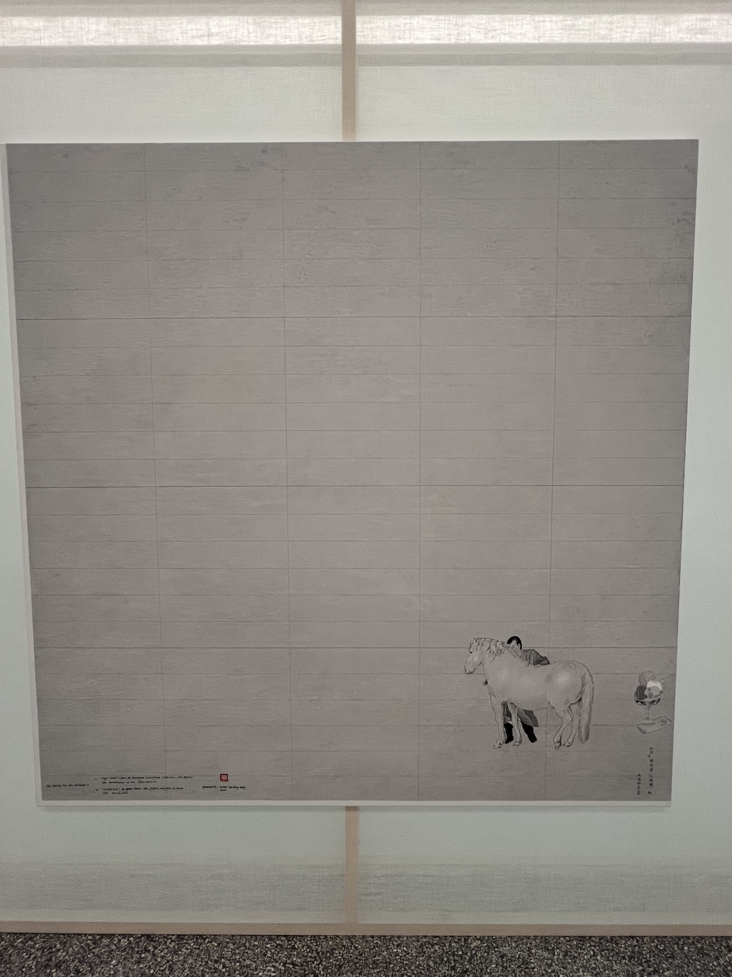 Gallery installation view of a large, mostly blank grey painting with faint horizontal ruled lines, featuring a small grayscale illustration of a person leaning against a horse in the lower right corner alongside handwritten annotations and a red artist's stamp.