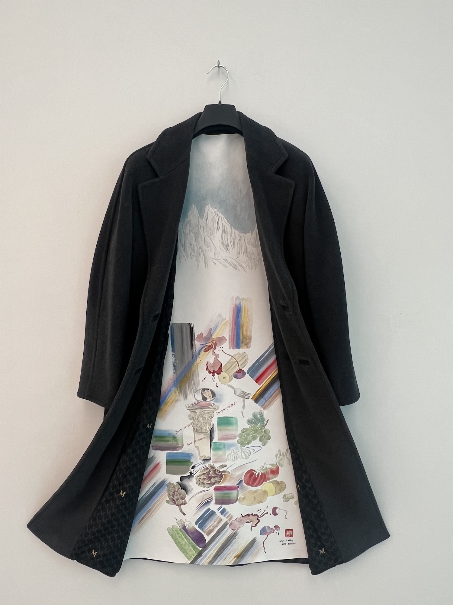 A black wool overcoat hangs open on a wall-mounted hook, revealing a painted lining that depicts a snow-capped mountain range at the top and a colorful still-life arrangement of vegetables, fruit, and abstract brushstrokes below, with handwritten text reading "So-so-sweet."