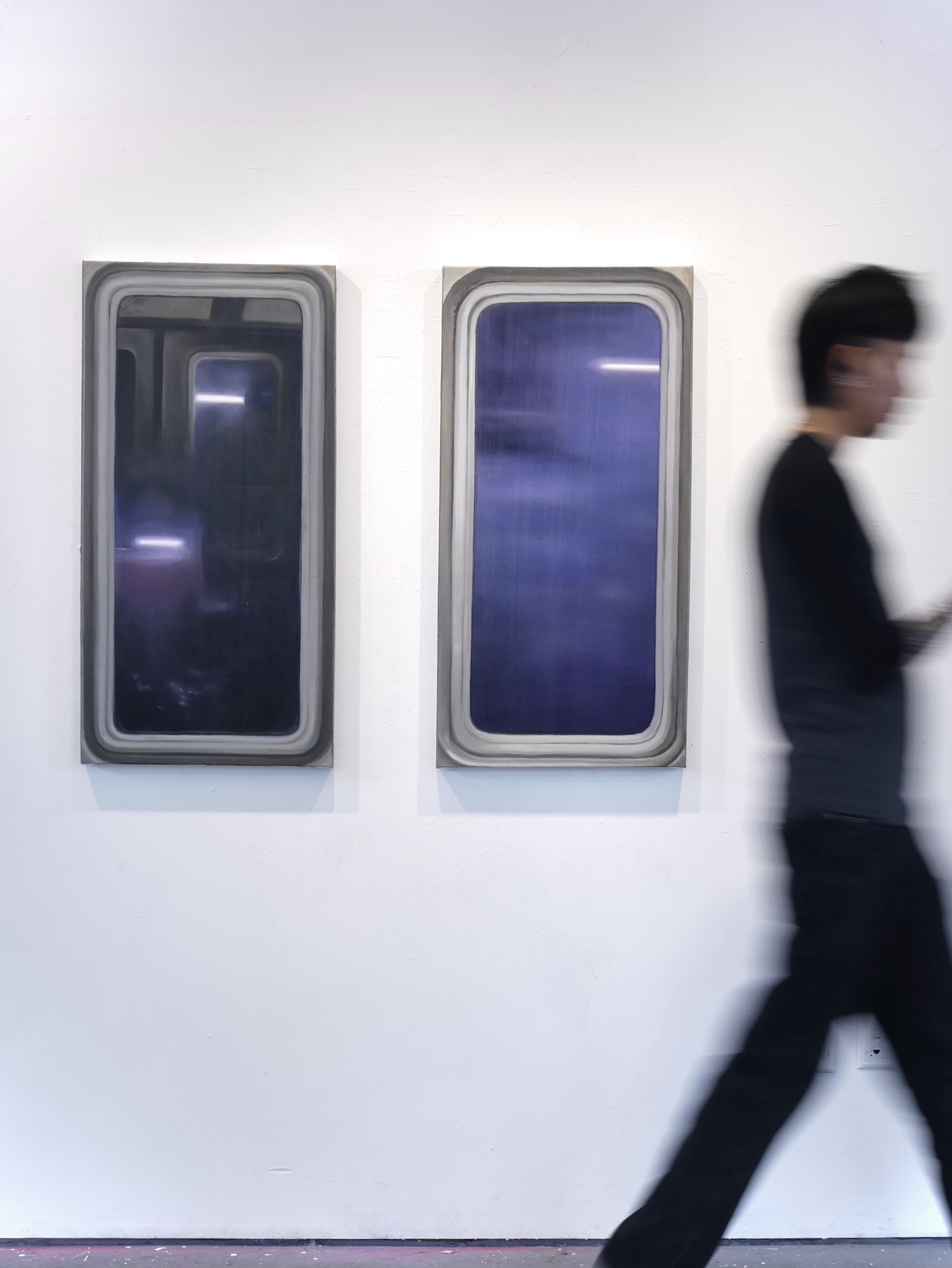 Two paintings resembling airplane windows with deep blue views hang side by side as a blurred gallery visitor walks past.