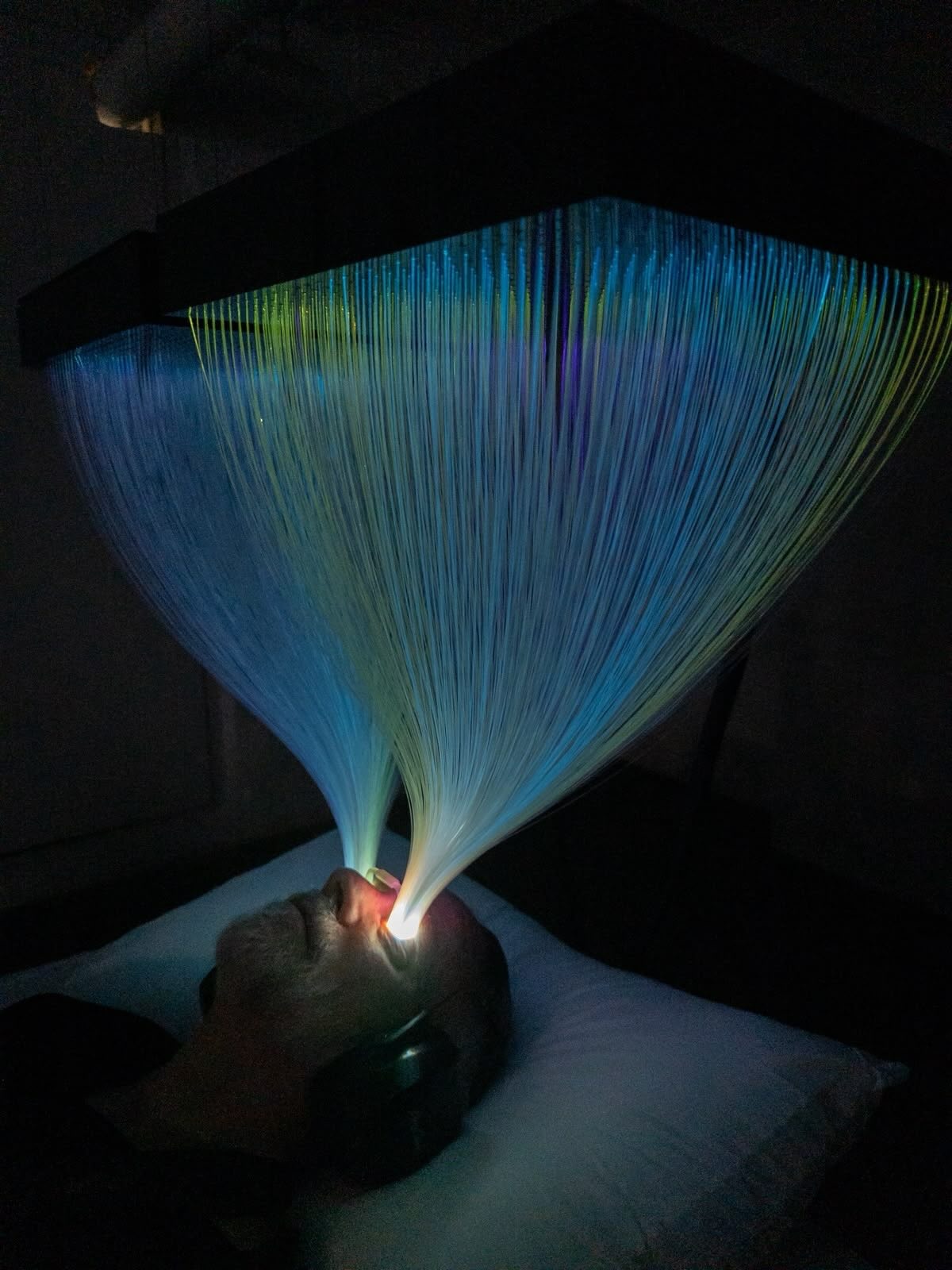 A person lies beneath a glowing fiber-optic installation, with illuminated strands appearing to flow into their eyes in a darkened room.