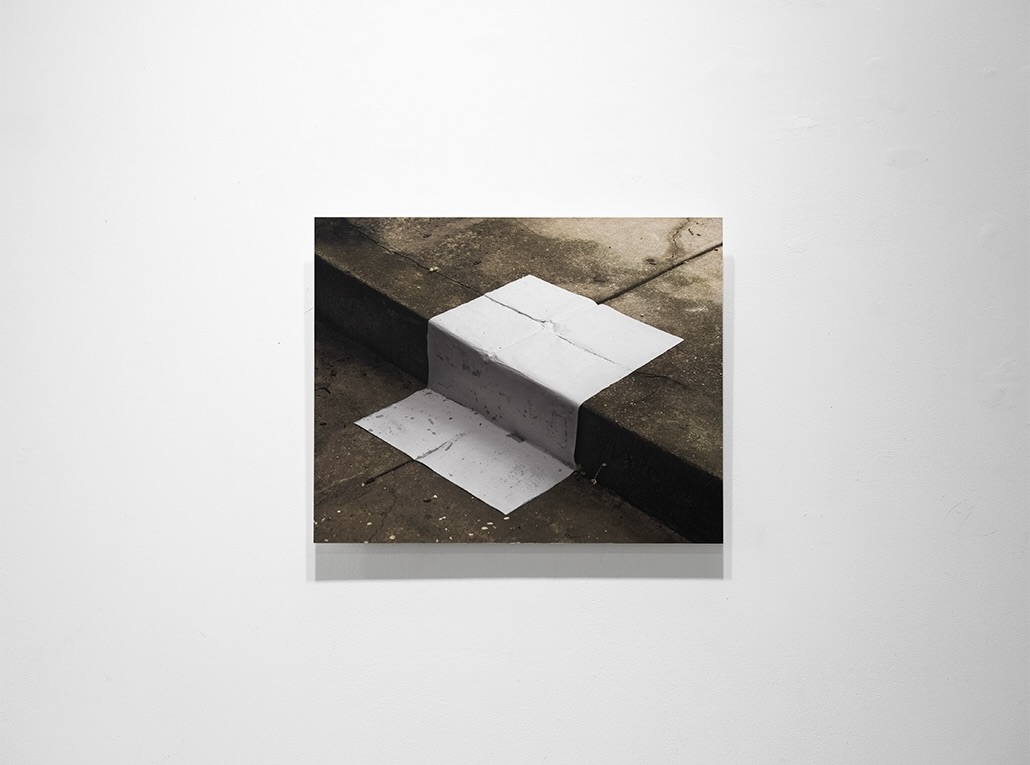 Photograph of a white paper or fabric form folded over a concrete curb edge, set against a weathered pavement backdrop.