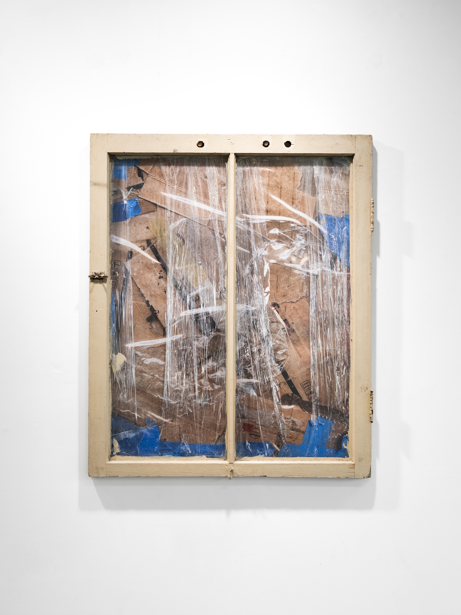 Two-pane wooden window frame covered in crumpled clear plastic wrap over layered cardboard and patches of blue tape, mounted on a gallery wall.