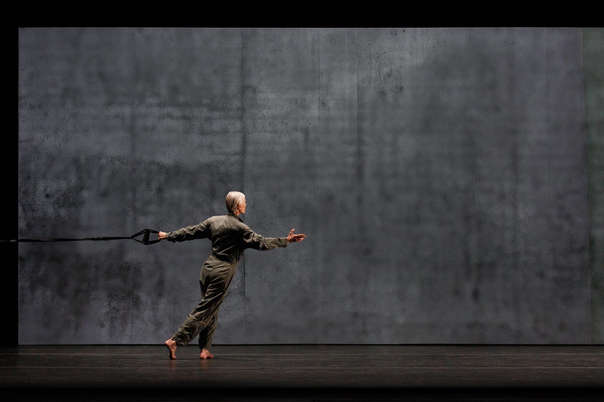 A silver-haired dancer in an olive jumpsuit leans forward on a dark stage, pulling against a harness strap, arms outstretched, against a bare concrete-textured backdrop.
