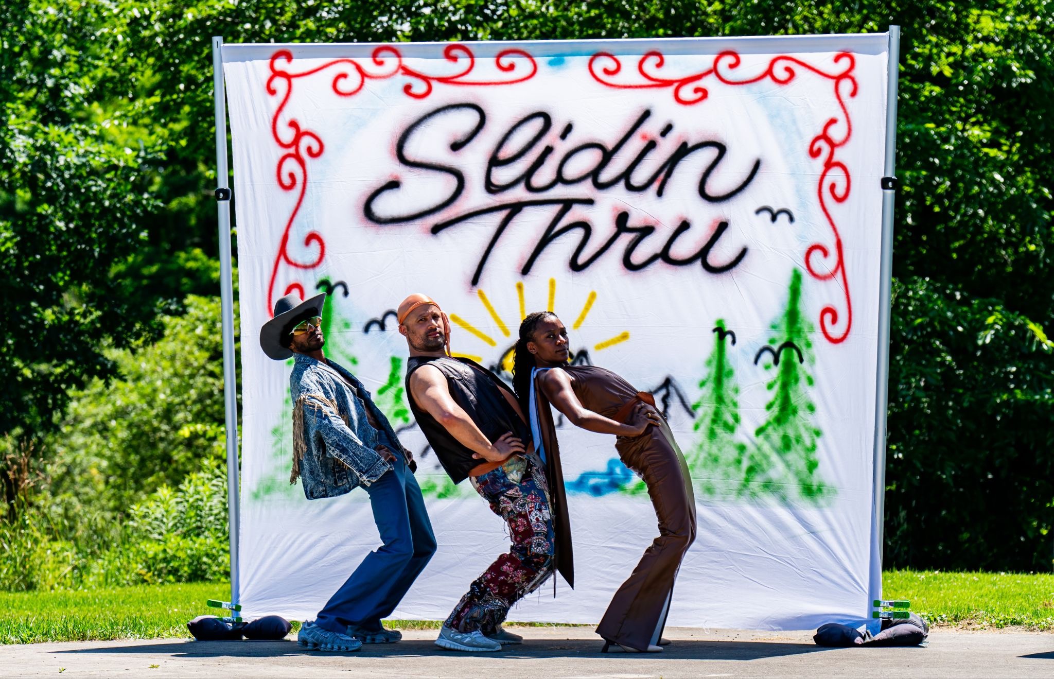 Three dancers pose dramatically in front of a hand-painted banner reading "Slidin Thru," decorated with red scrollwork, a sun, mountains, and trees. Outdoors on a sunny day, surrounded by greenery.