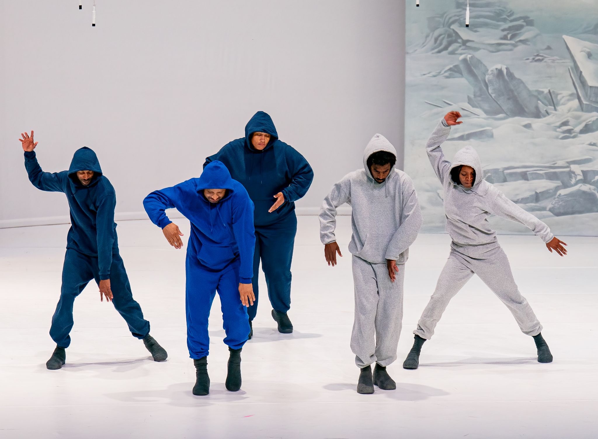 Five dancers in blue and gray hoodies perform in dynamic, angular poses on a white stage, with a painted icy mountain backdrop visible at right.