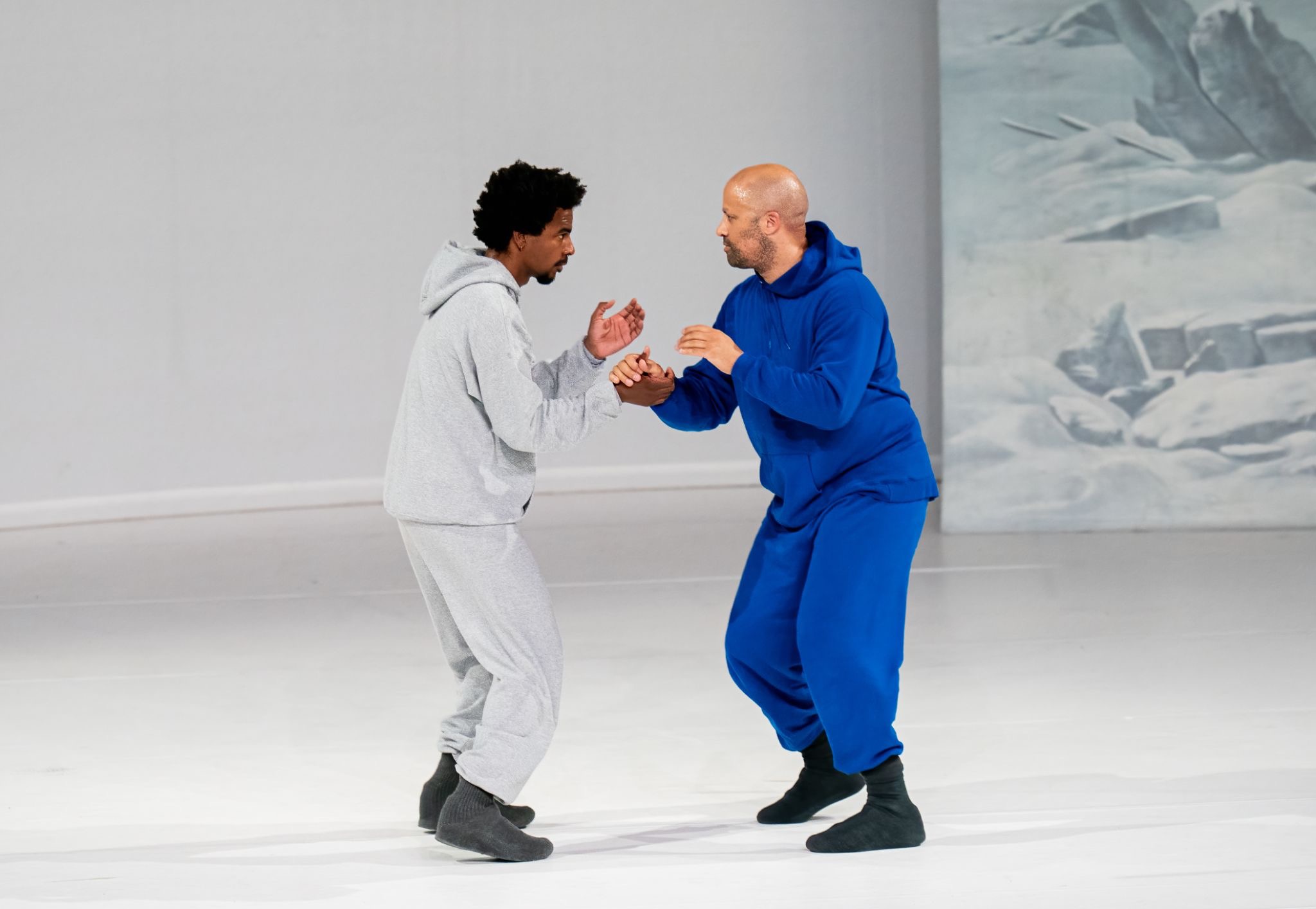 Two dancers in gray and blue sweatsuits face each other in a crouched stance, clasping hands, on a white stage with an icy landscape backdrop.
