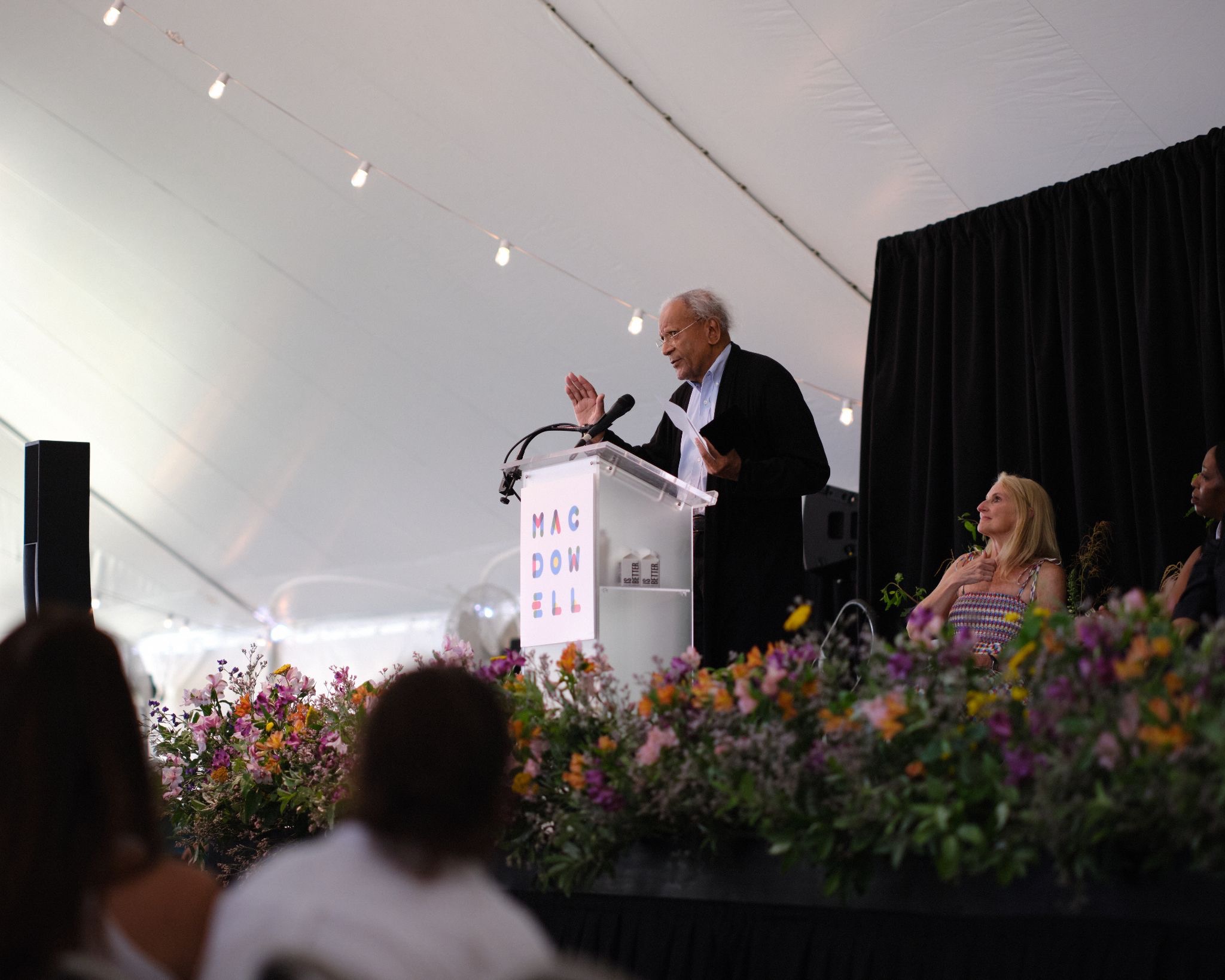 A man in a dark cardigan speaks at a podium reading "MacDowell" under a white event tent, with a woman seated nearby and colorful flower arrangements in the foreground.