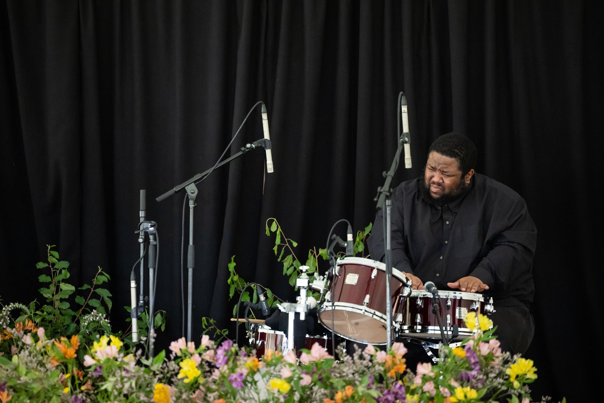 A drummer in a black shirt plays a set of drums on a stage framed by a black curtain, condenser microphones, and colorful flowers in the foreground.