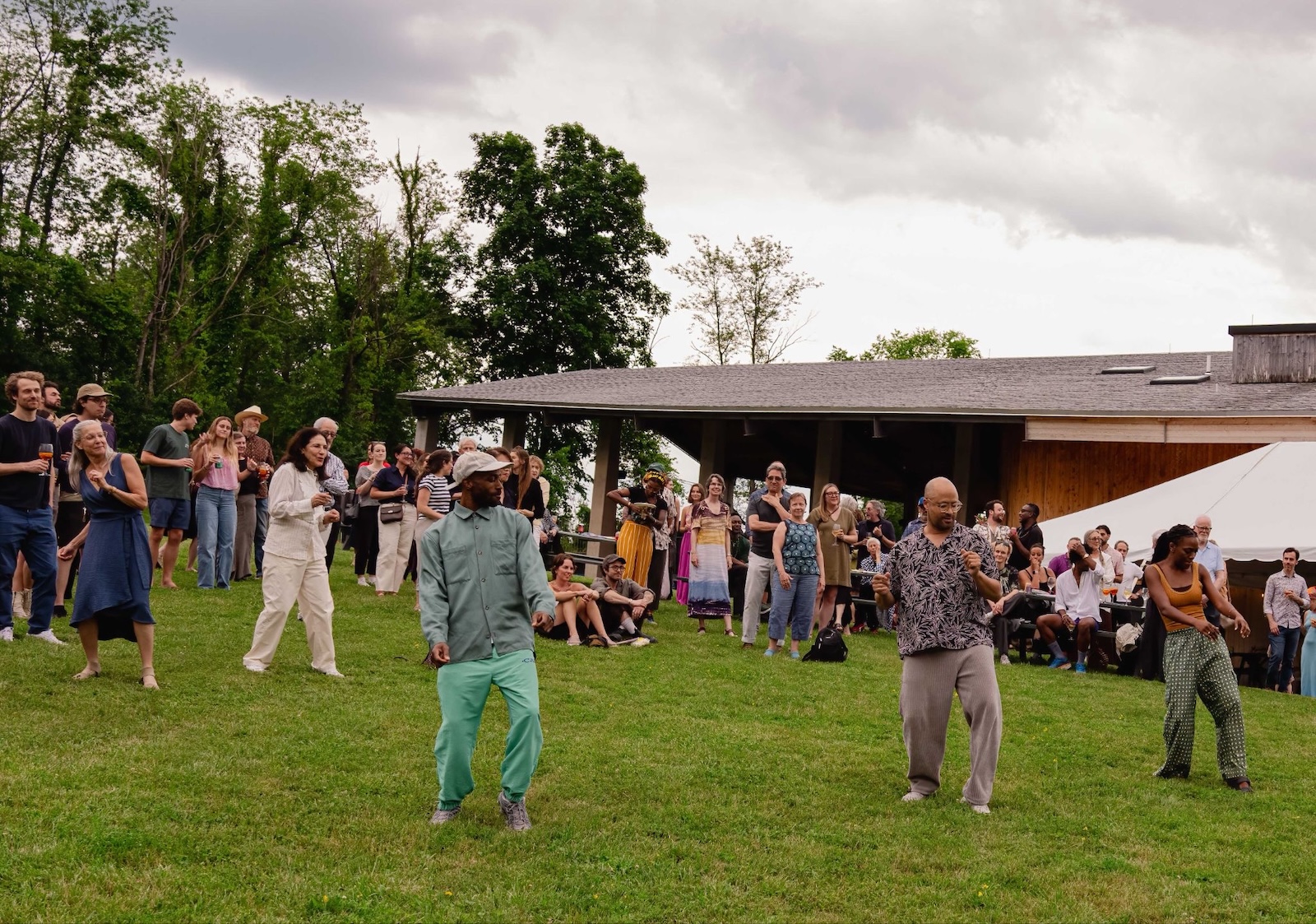 A crowd gathers on a lawn under a cloudy sky as several people dance in the foreground, with a pavilion tent and audience members watching in the background.