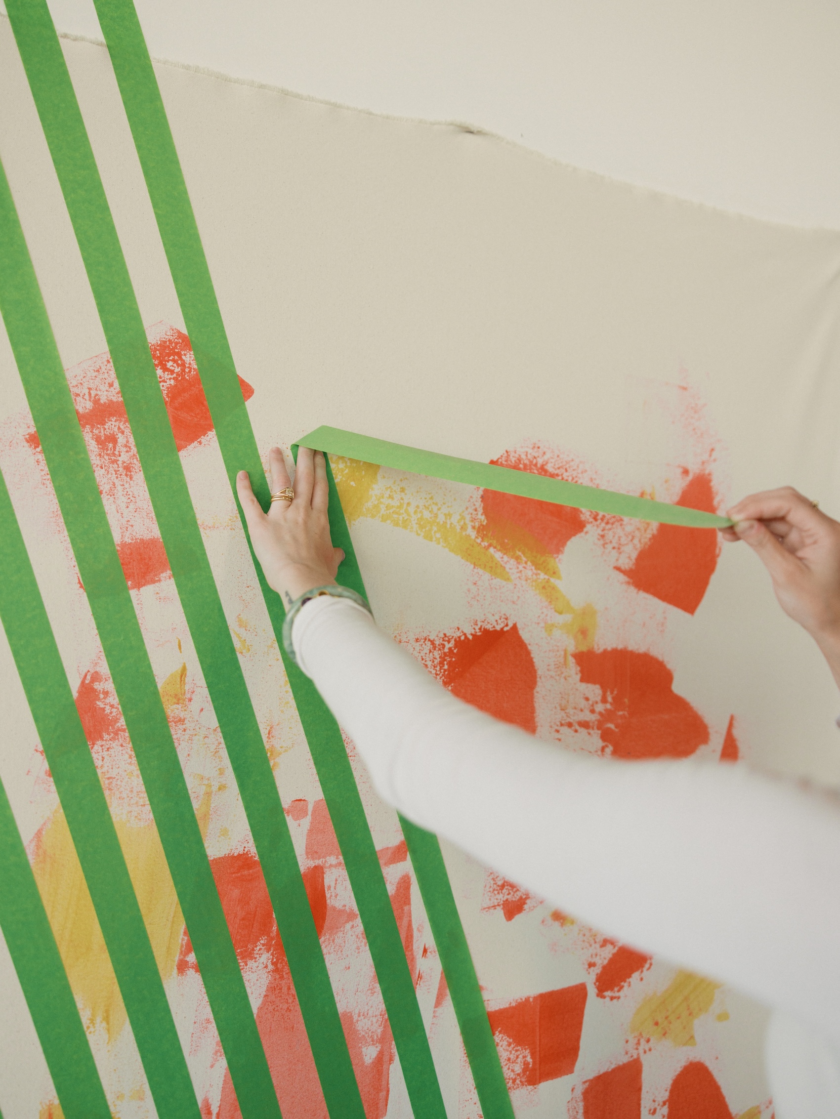 Close-up of an artist's hands pulling green painter's tape off a canvas, revealing an abstract pattern of orange and yellow sprayed and layered shapes underneath.