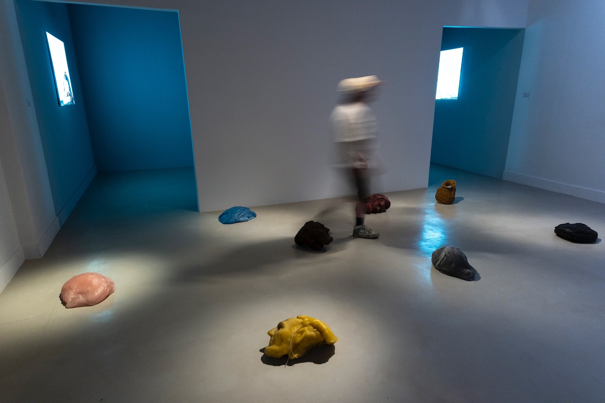 Gallery installation view with blurred visitor walking past sculptural, blob-like ceramic forms in varied colors (pink, blue, yellow, black, ochre) scattered across the floor, with video screens visible in blue side alcoves.
