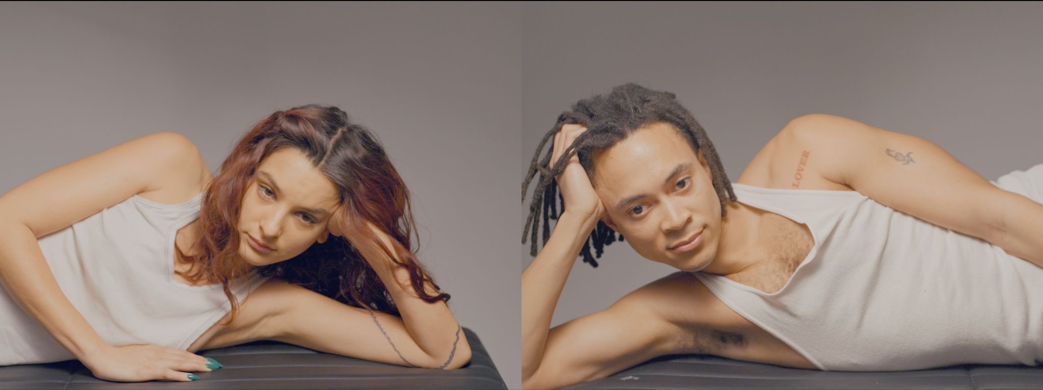 Split-screen portrait of two people in white tank tops reclining and gazing at the camera, one with long red-brown hair, the other with gray dreadlocks and a "LOVER" arm tattoo, against a neutral gray background.