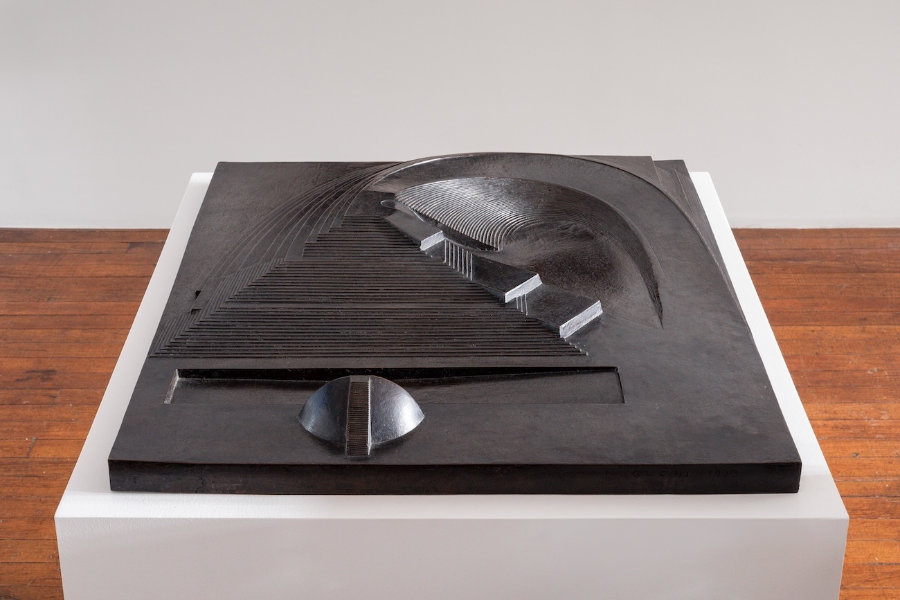 Dark bronze relief sculpture on a white pedestal depicting an abstracted stepped ramp and curved slide form, resembling an aerial view of a playground or slide structure.