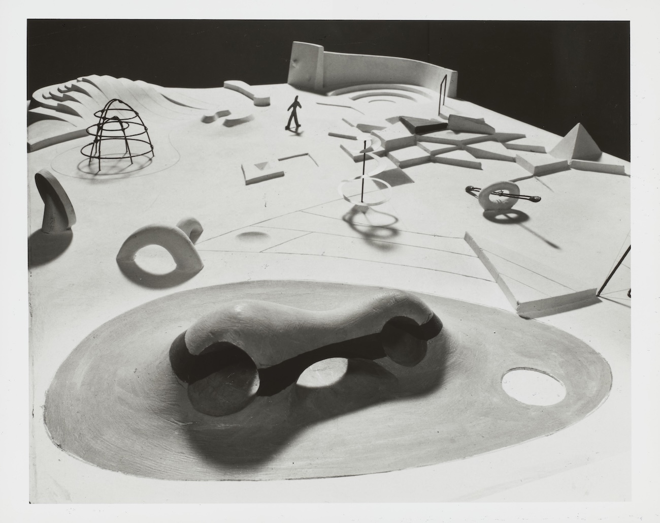 Black-and-white photograph of a scale model for a playground design, showing a sculpted landscape with abstract climbing structures, a dome-shaped jungle gym, geometric shapes, and a tiny figure for scale; a curved tunnel-like form sits in the foreground.