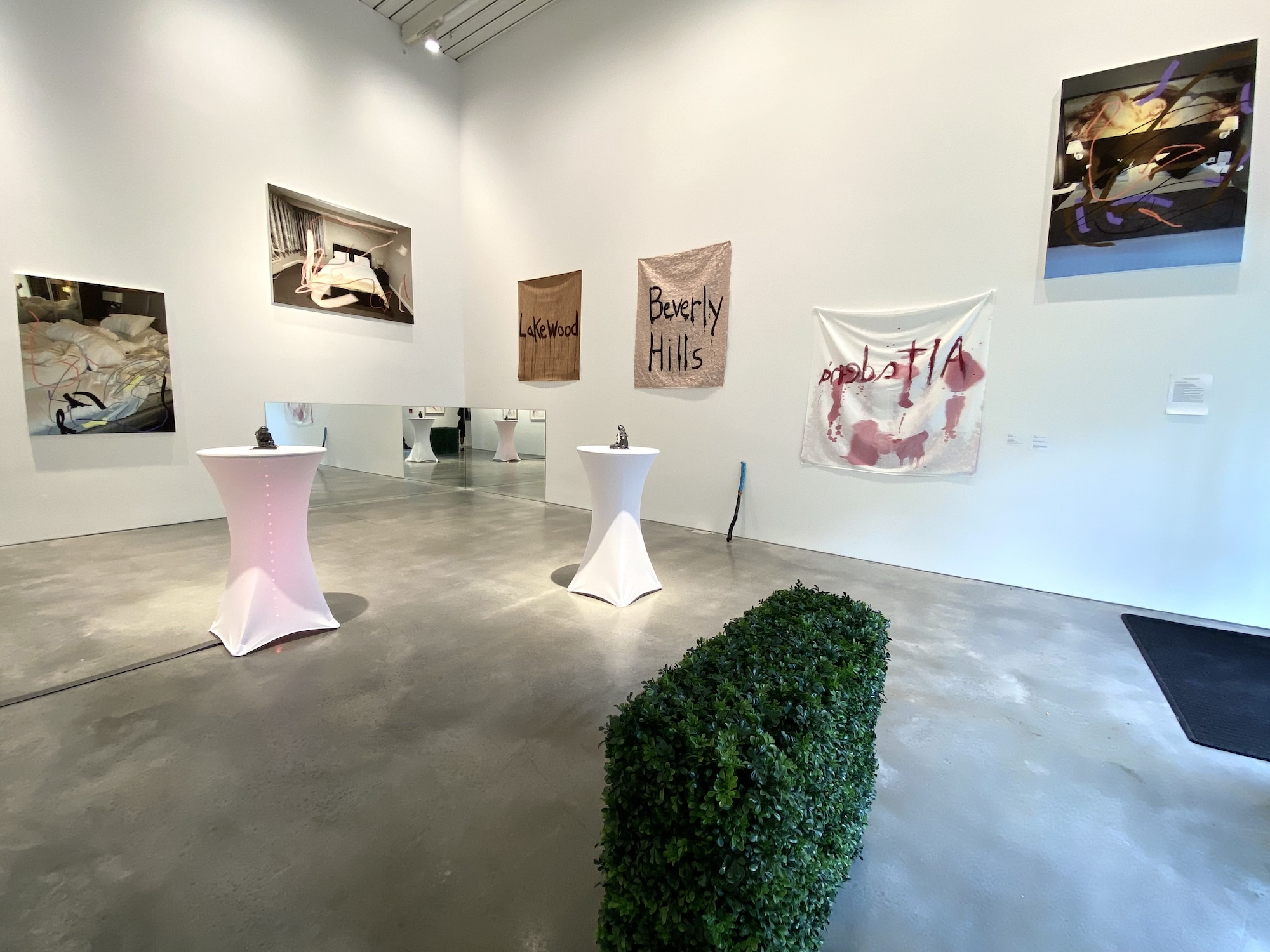 Gallery installation view with white walls hung with photographs of tousled hotel beds, fabric banners reading "Lakewood" and "Beverly Hills," a blood-red painted textile banner, and small sculptures on pedestal tables, with a boxwood hedge and mirrored wall in the foreground.