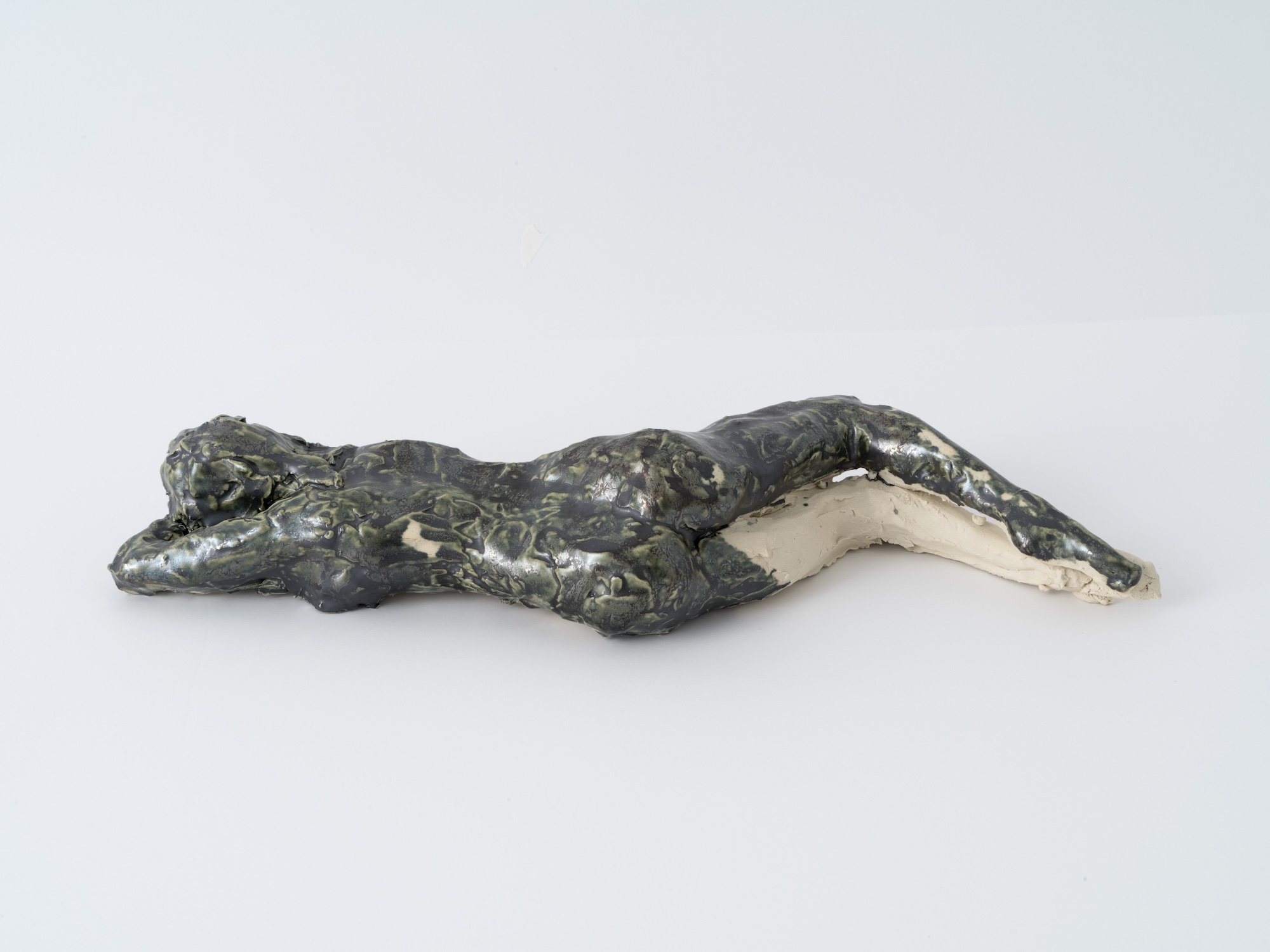 Reclining nude figure ceramic sculpture with a mottled black-and-green glaze over unglazed white clay, lying on its stomach with head resting on folded arms.