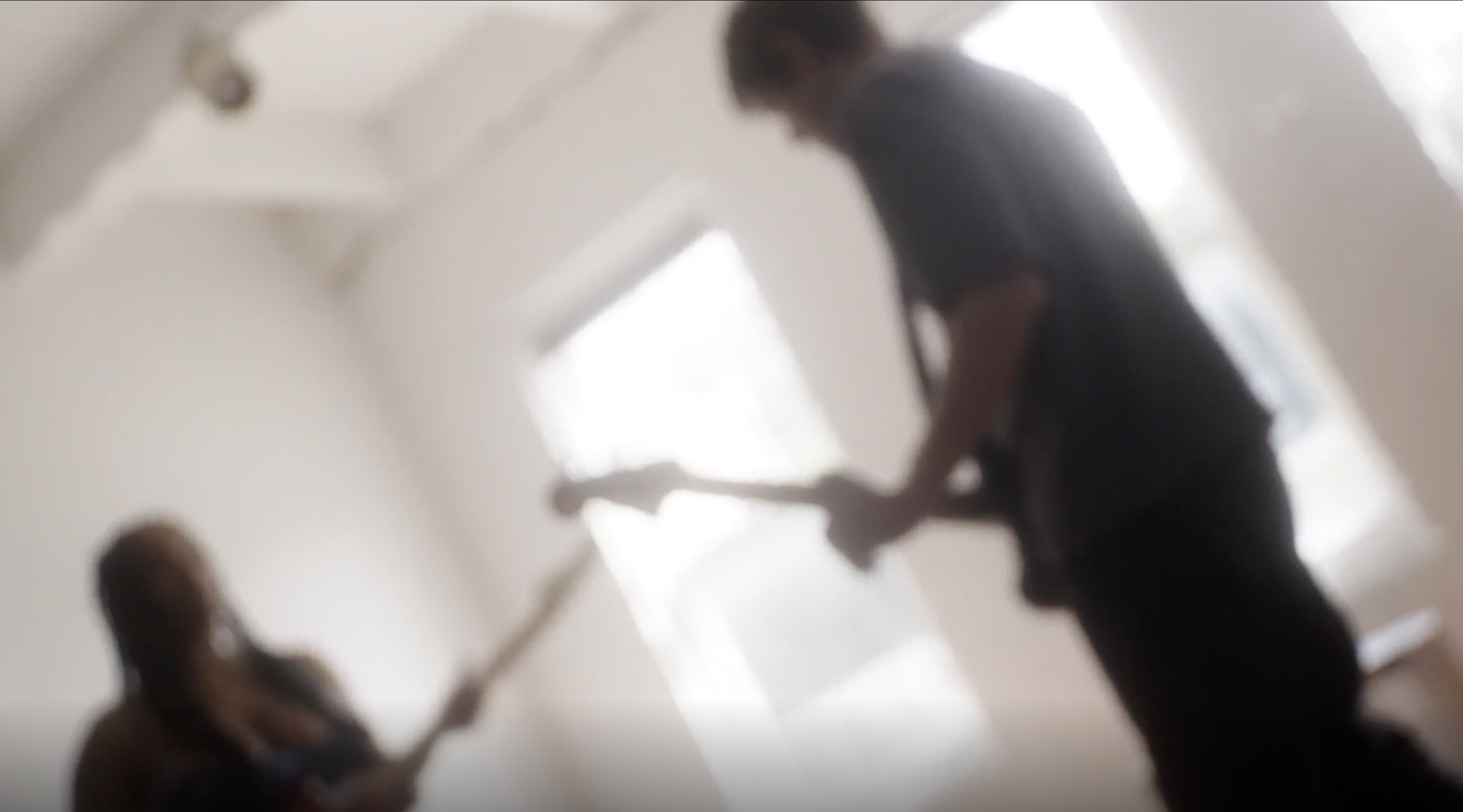Blurred, low-angle photograph of two figures playing electric guitars in a bright, white-walled room with tall windows, motion blur creating a hazy, dreamlike effect.
