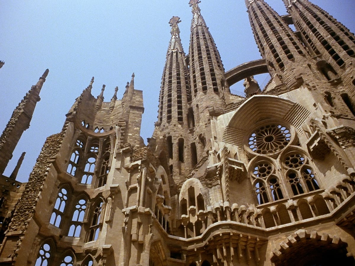 Low-angle view of the Sagrada Família's ornate Gothic spires in Barcelona, with intricately carved stonework, a rose window, and tall pinnacles against a clear blue sky.