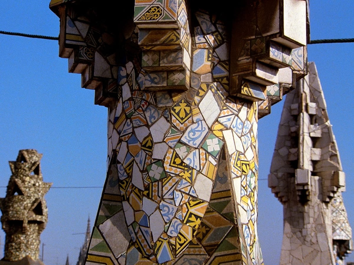 Close-up of a colorful mosaic-tiled chimney with broken ceramic fragments in blue, yellow, green, and white patterns, set against a clear sky with similar decorative chimneys nearby.