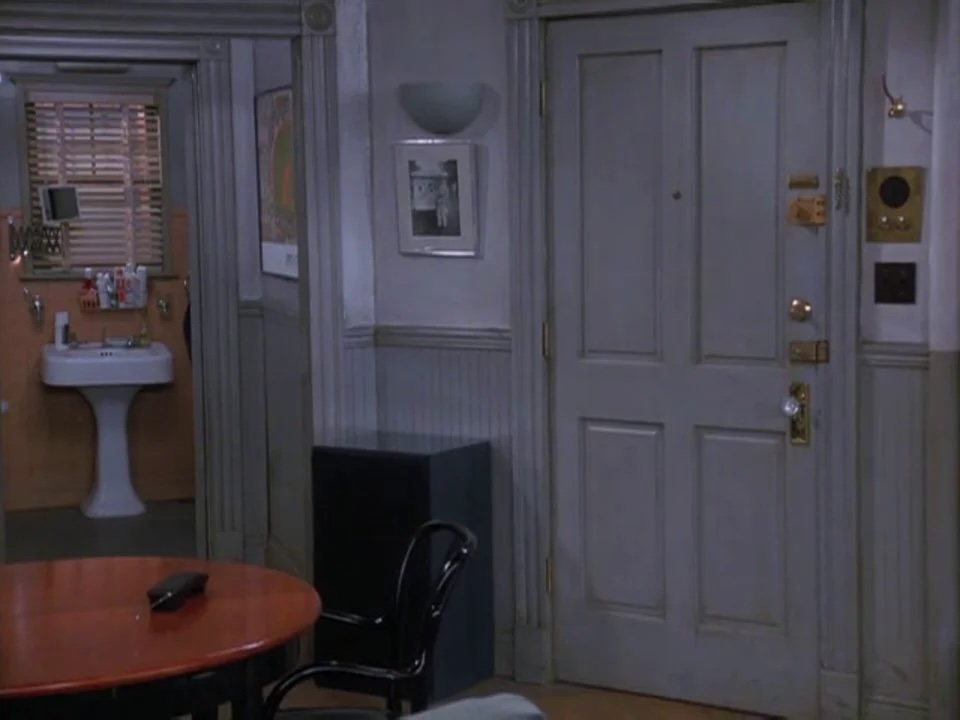 Sitcom apartment interior with a white paneled door, gold hardware, an intercom panel, a black bistro chair beside a round orange table, and a bathroom sink visible through an open doorway.