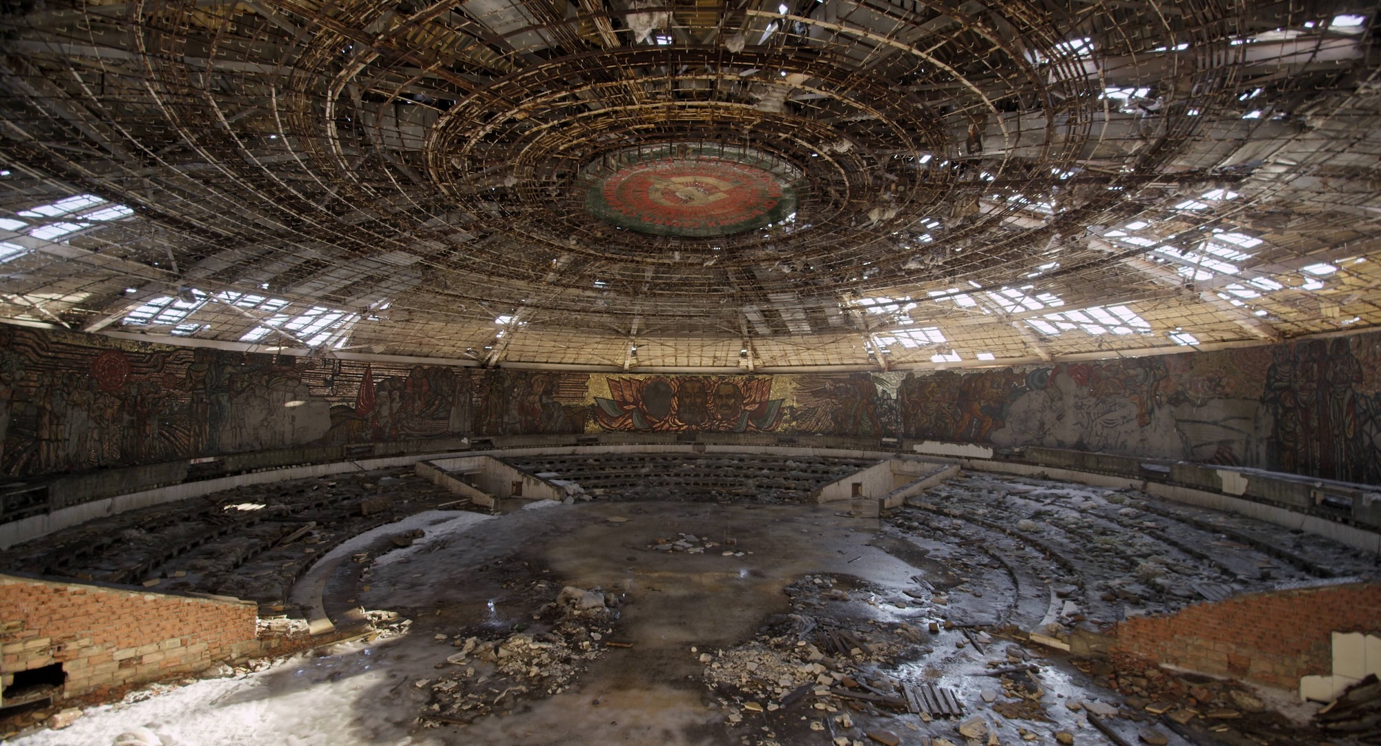 Interior of a decaying, saucer-shaped Soviet-era monument (Buzludzha Monument) with a domed, ribbed ceiling open to the sky, faded mosaic murals along the curved walls, and rubble scattered across the floor.