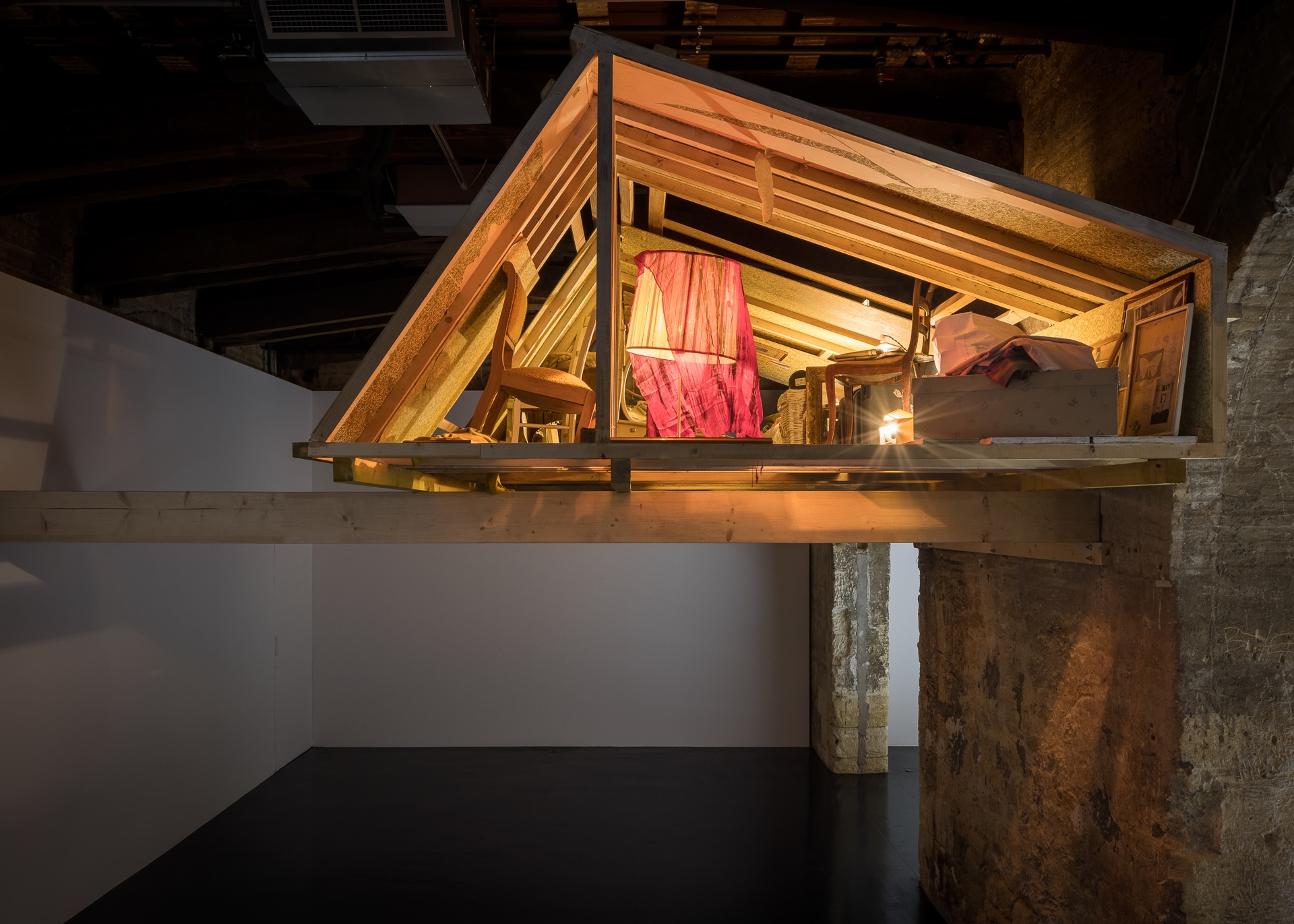 A house-like attic structure is suspended above the floor inside a dark gallery. Its exposed wooden rafters frame chairs, boxes, baskets, fabric, and lamps glowing warmly within the elevated, cluttered interior.