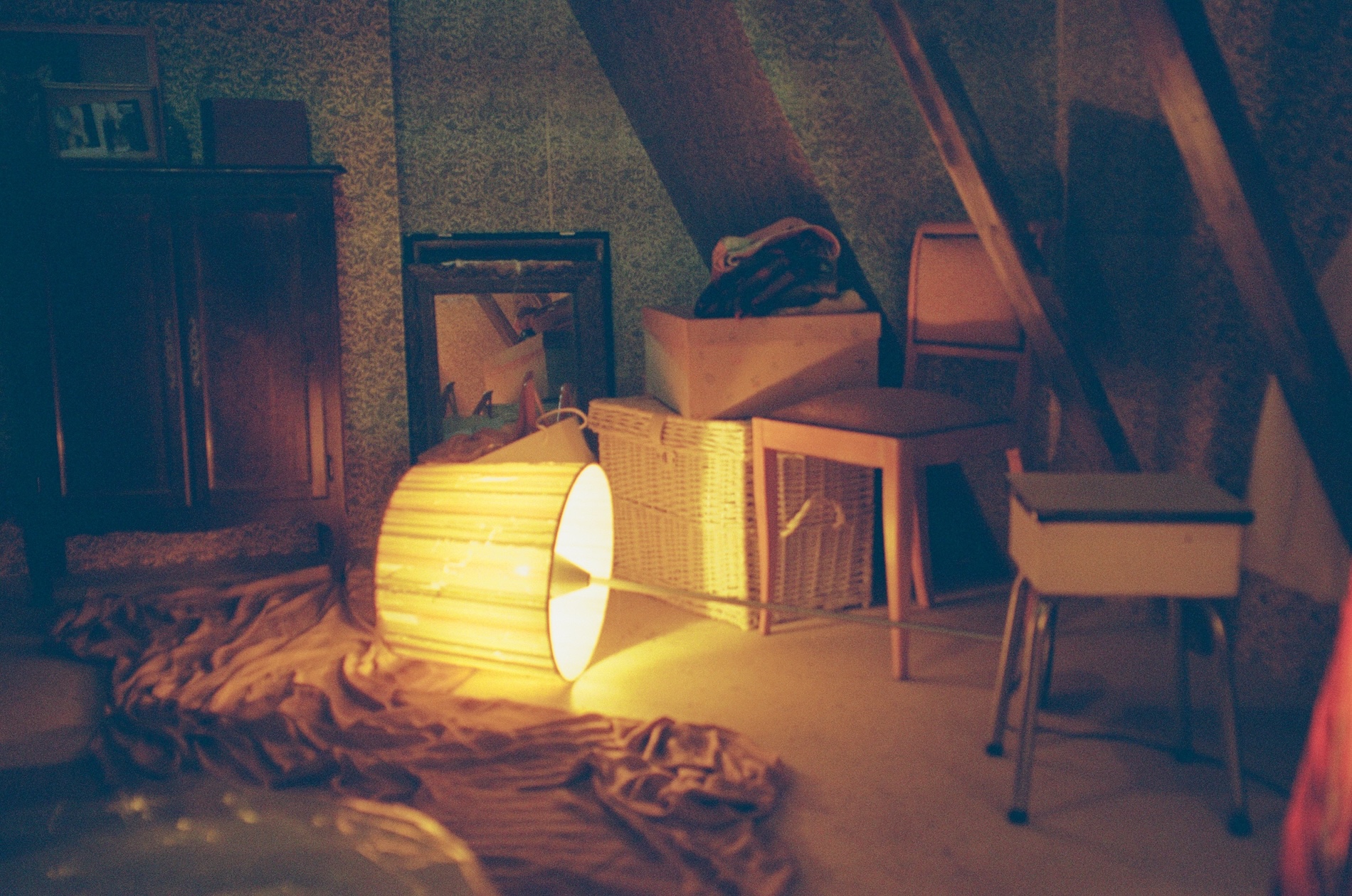 Dimly lit attic interior filled with mismatched furniture, baskets, fabric, and stored belongings. A lampshade lies on its side on the floor, casting a warm yellow glow across rumpled fabric.