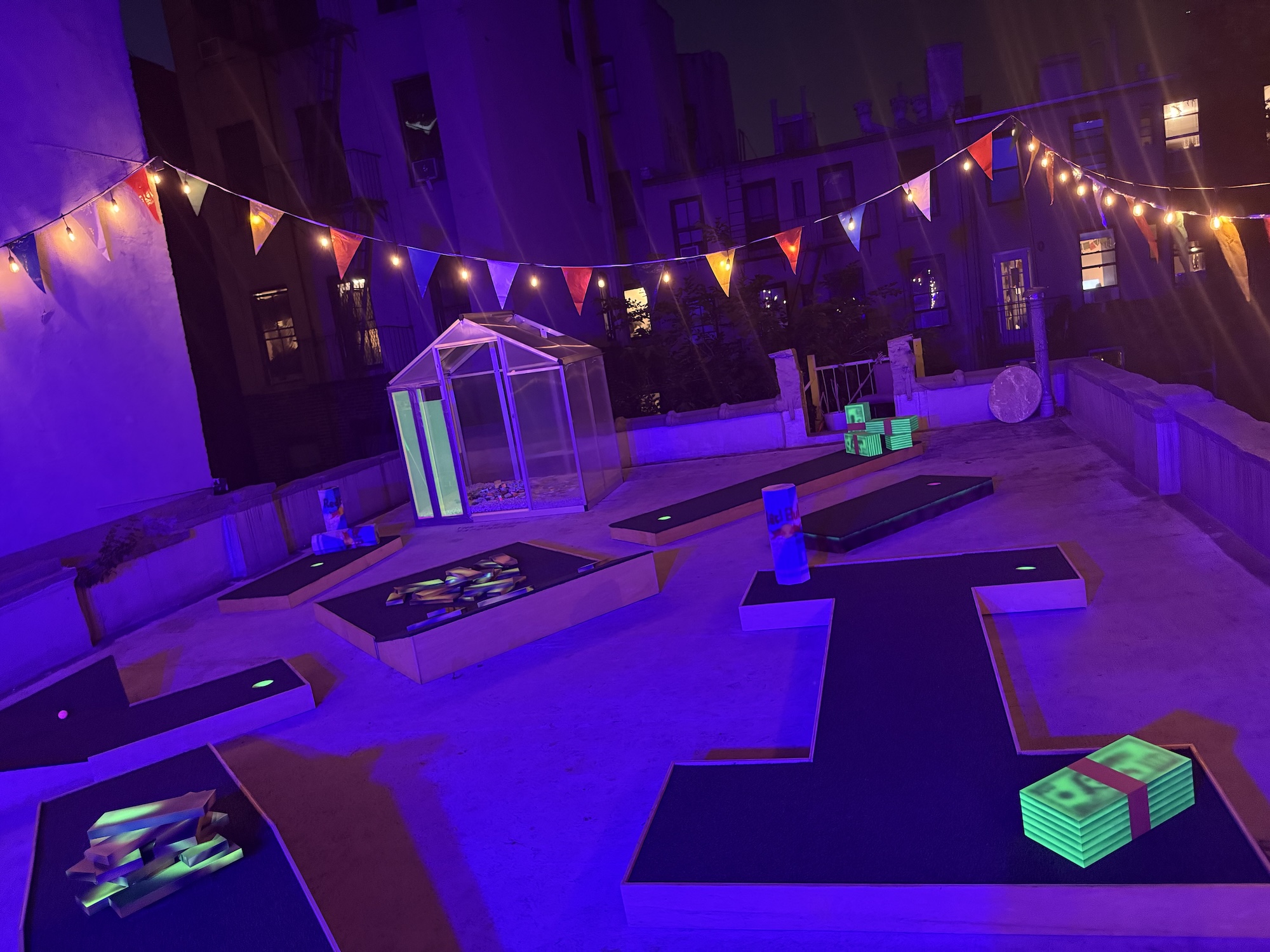 Rooftop miniature-golf installation illuminated by purple and ultraviolet light, with glowing sculptures, string lights, and colorful flags.
