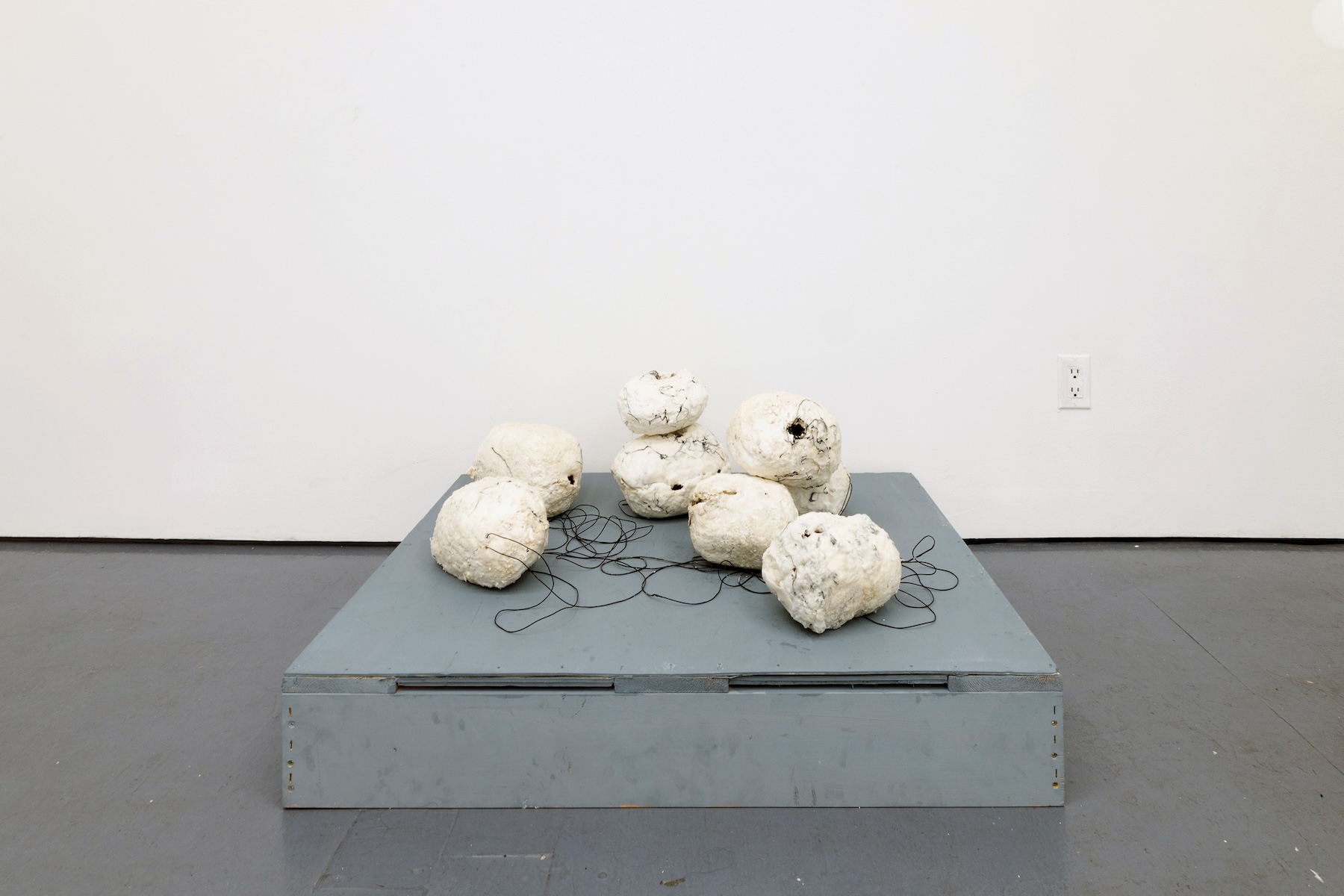 Installation of rough, white spherical sculptures connected by loose black wires, arranged on a low gray platform in a gallery.