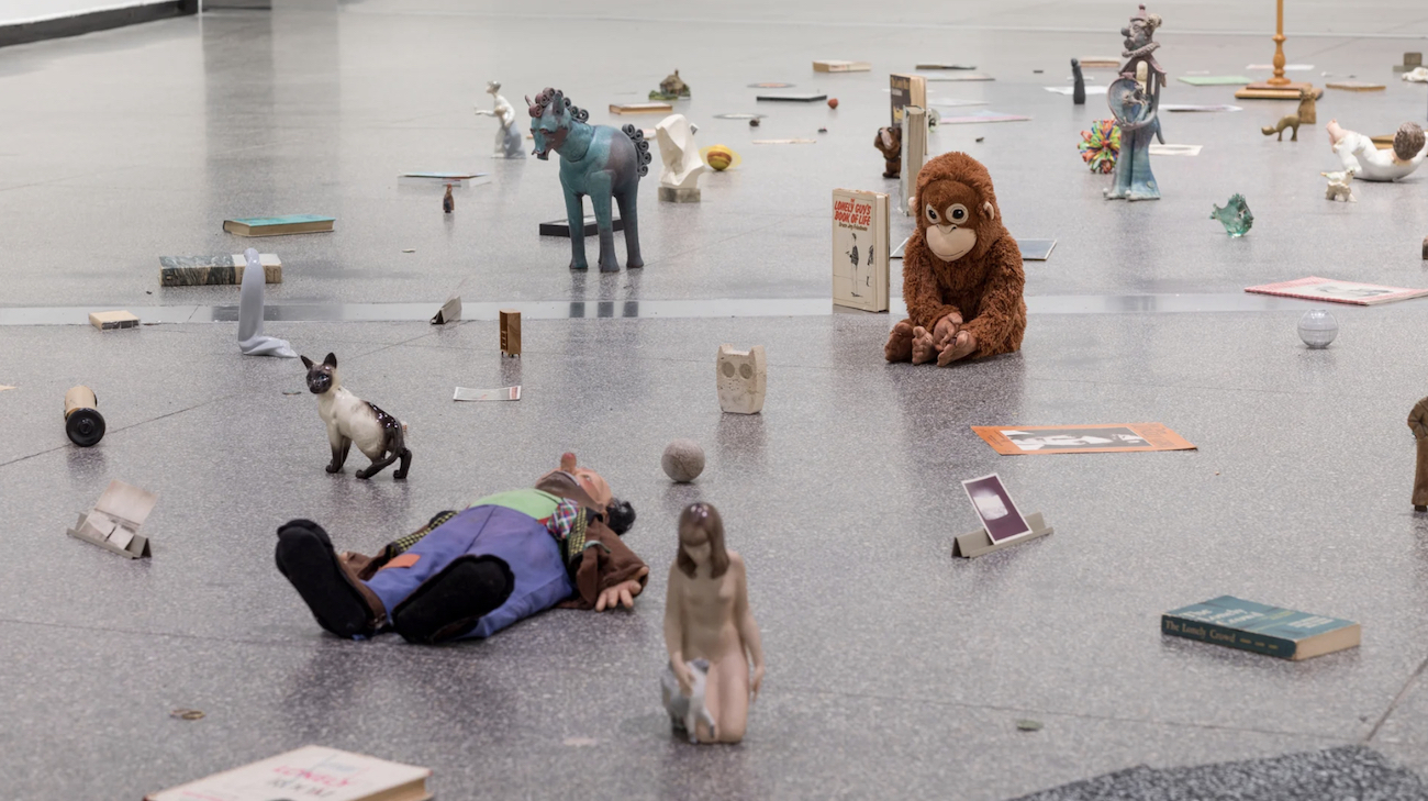 Books, figurines, toys, photographs, and other small objects are loosely scattered across a gallery floor.