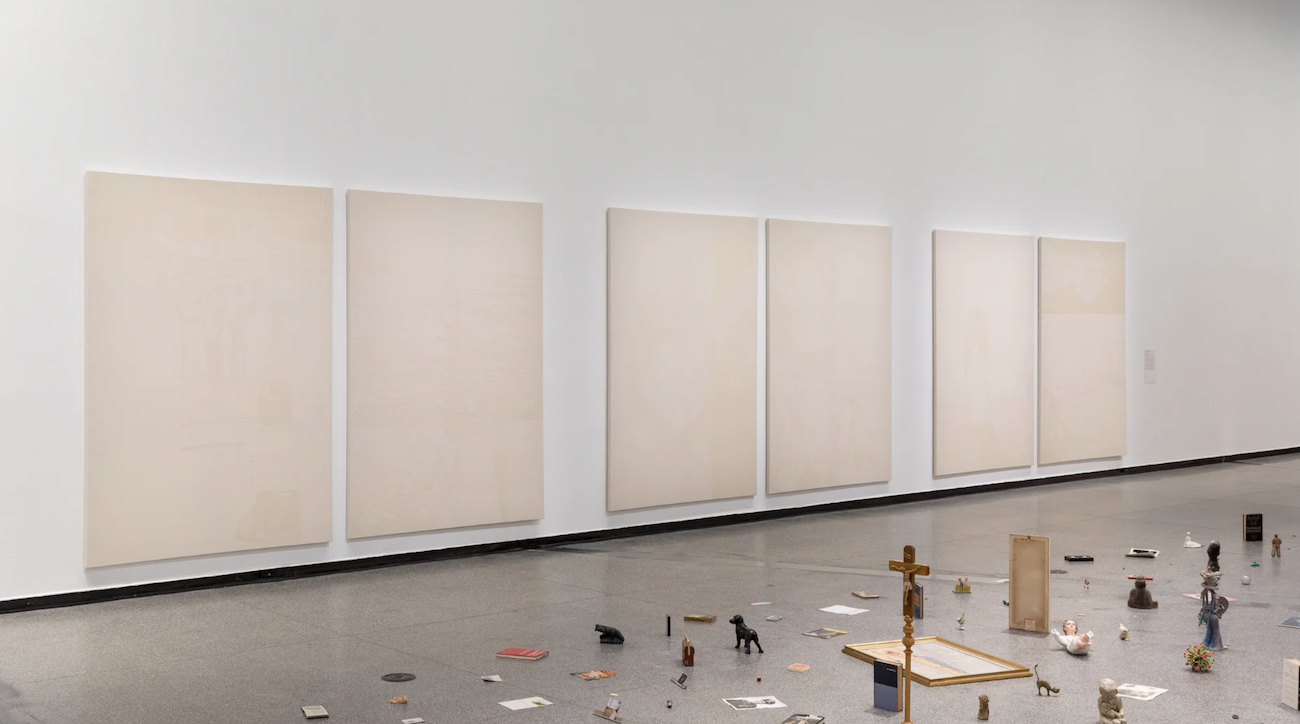 Six pale, nearly monochromatic rectangular paintings hang in pairs above an installation of scattered objects.