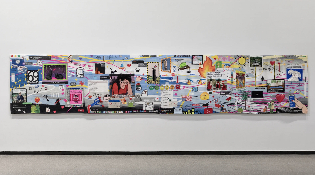 A long embroidered textile resembling a chaotic digital desktop hangs across a white gallery wall.