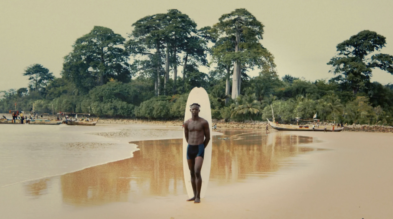 A lone surfer carrying a white surfboard walks along a beach backed by dense trees.