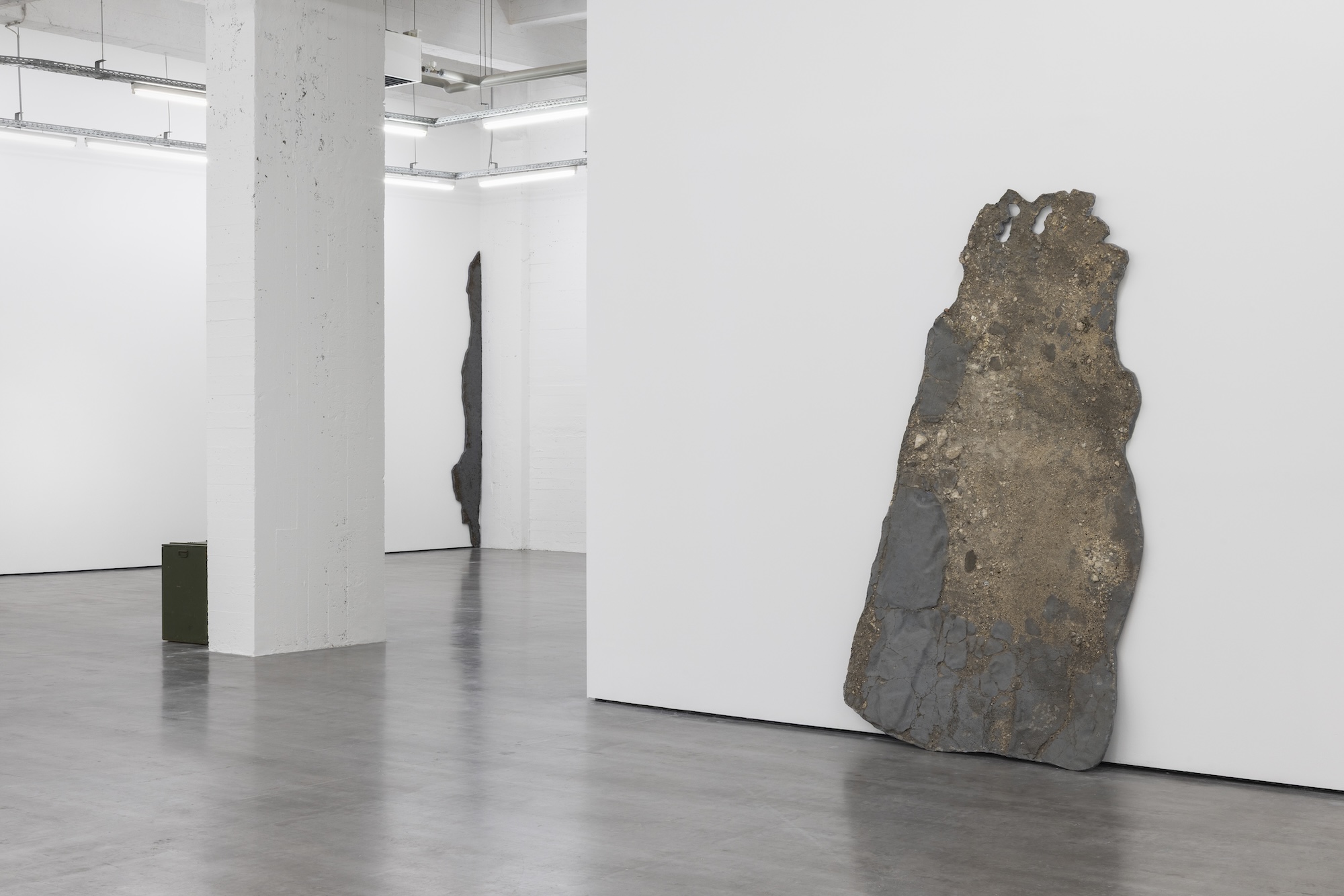 Large irregular slabs of earth- and asphalt-like material lean against white gallery walls, with a small green metal box positioned near a central column.