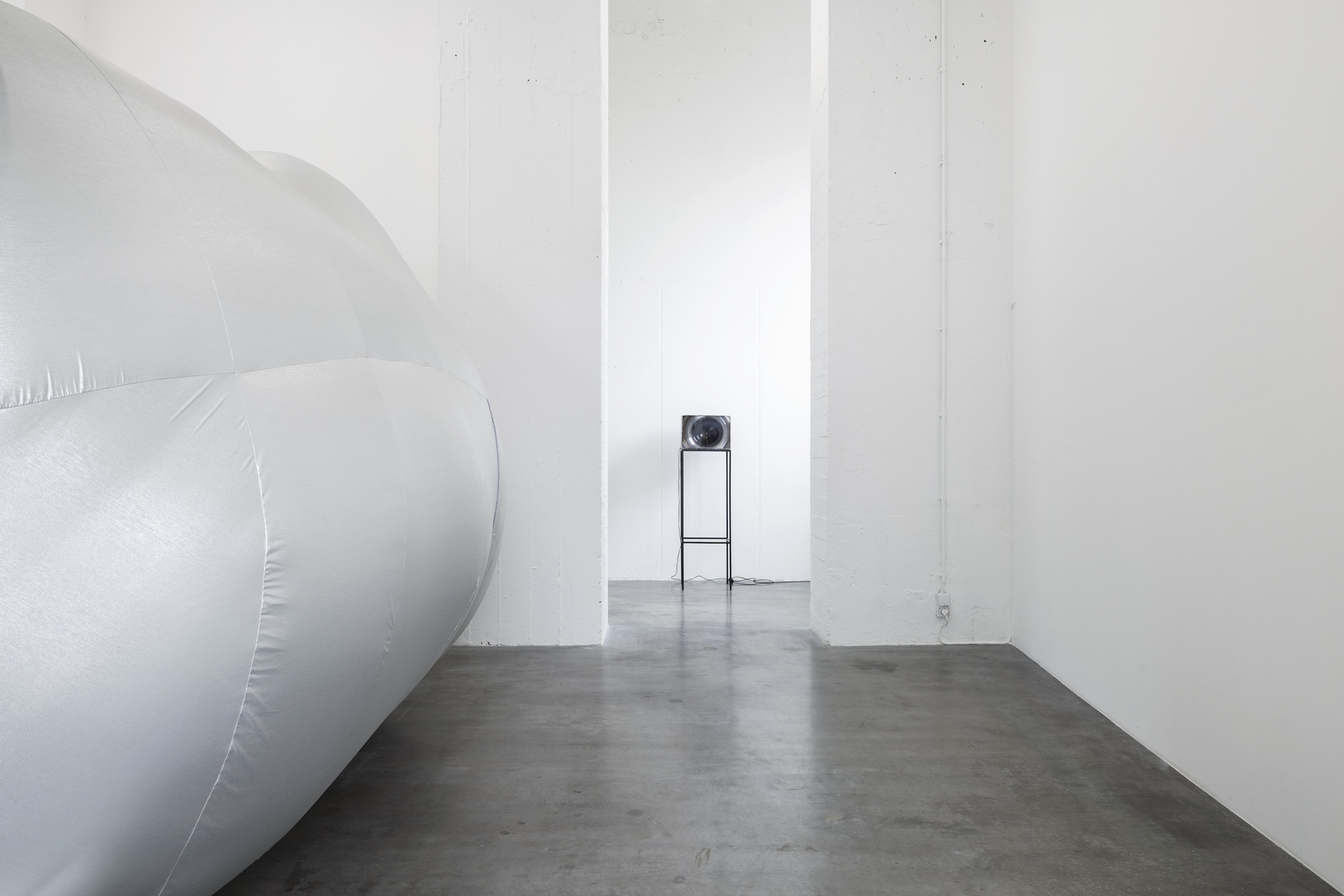 A monumental silver inflatable form fills the left side of a white gallery space, with a small circular object mounted on a tall black stand visible through an opening.