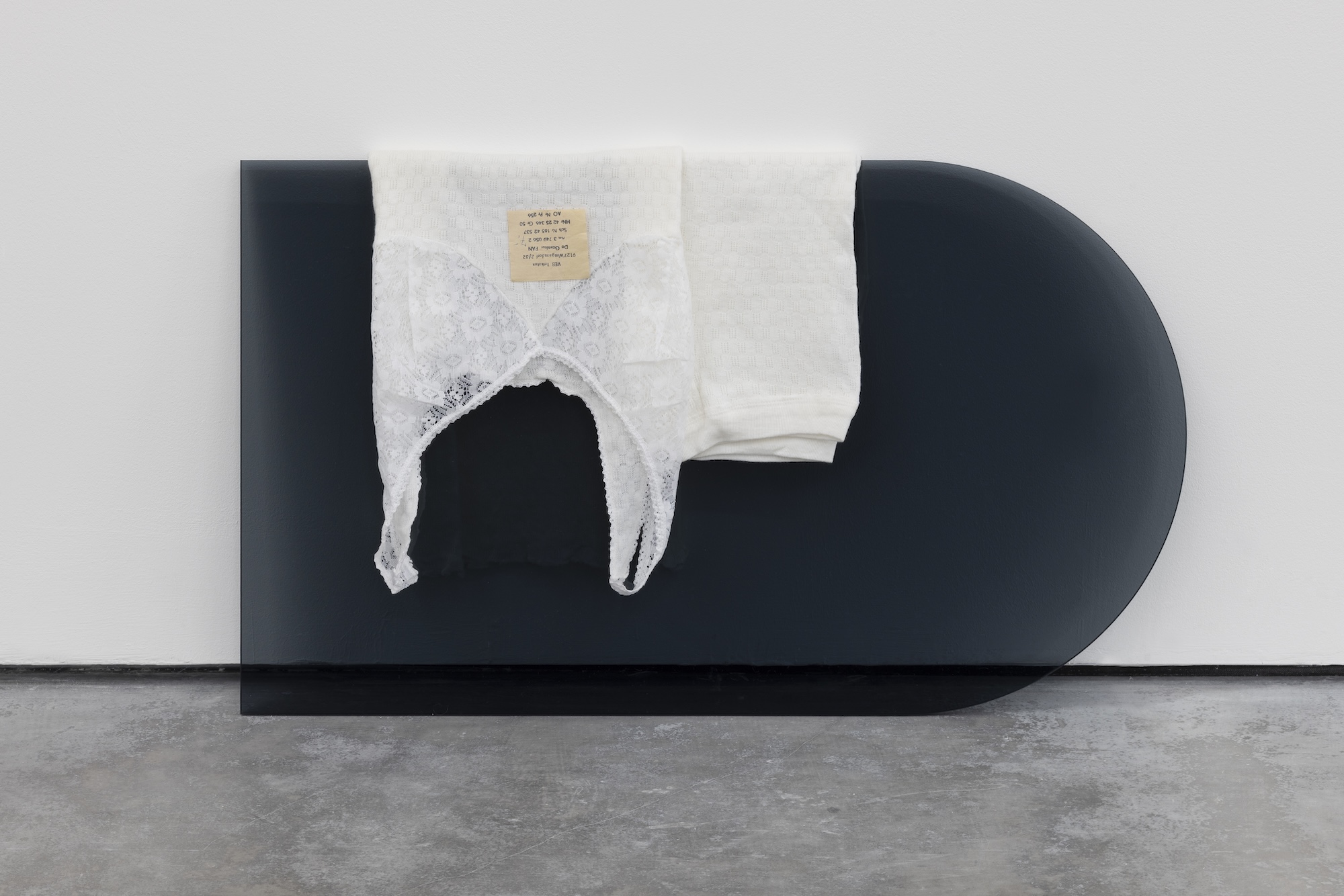 White lace underwear and folded white fabric drape over a low, black, rounded rectangular structure against a gallery wall.